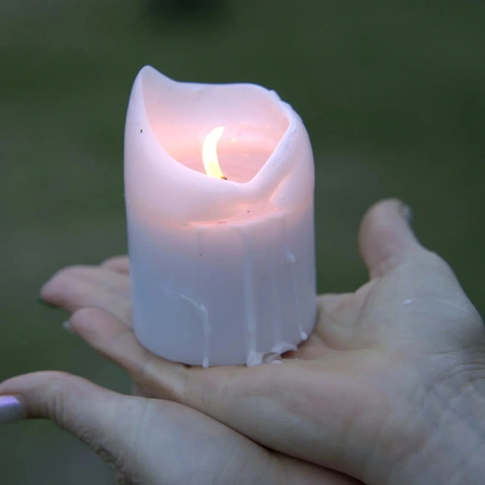 A hand holding a white candle with a flame inside, partially melted, with a blurry green background.