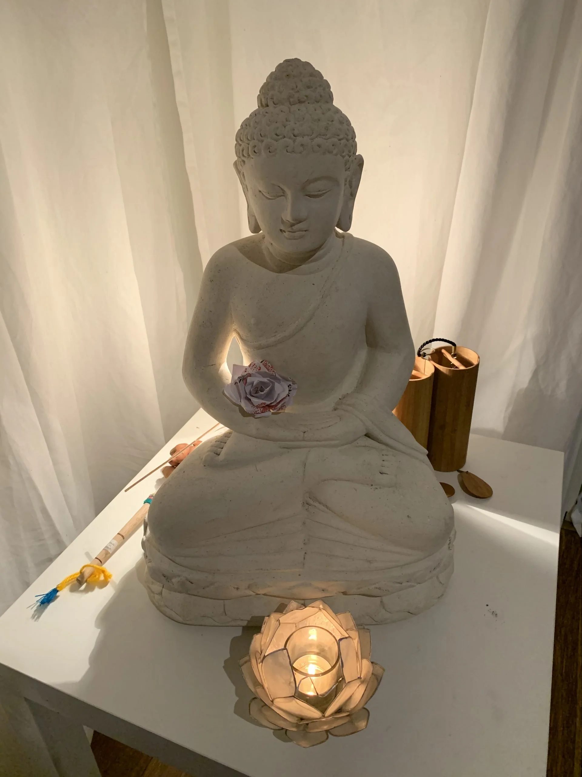 White Buddha statue with closed eyes, seated in meditative pose, holding a folded paper flower, on a white table with traditional wooden lanterns and a candle holder, set against white curtains.