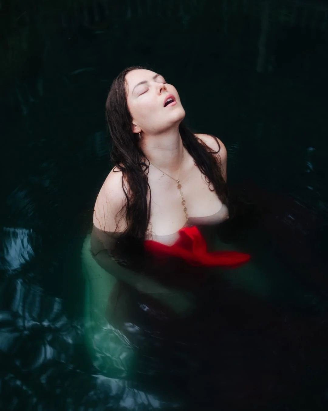 A woman with wet hair and closed eyes, partially submerged in dark water, wearing a red top and a gold necklace.