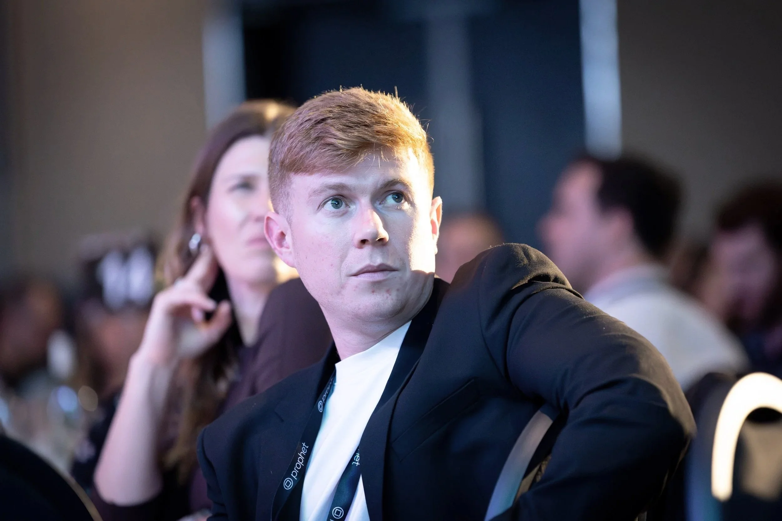 A young man with short, reddish hair takes a selfie at a conference, wearing a black blazer and white shirt, with other attendees blurred in the background.