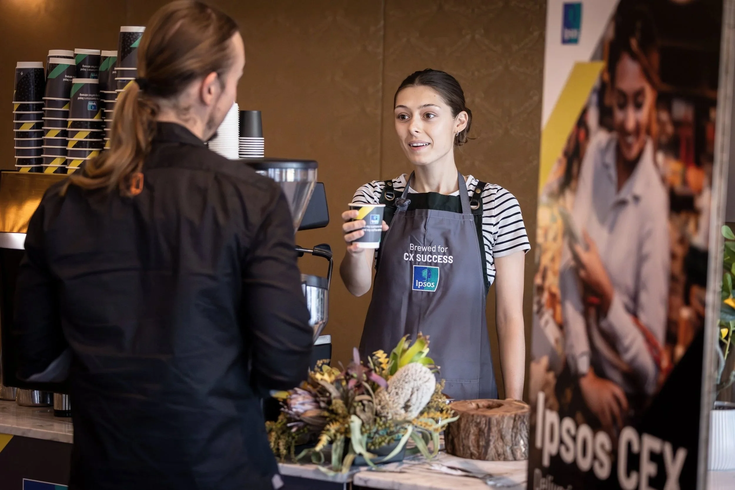 A young woman working at a coffee stand, talking to a customer with her hair tied back, wearing a striped shirt and gray apron with the text 'Brewed for CX Success.' There are coffee cups stacked behind her, and a flower arrangement on the counter in front of her.