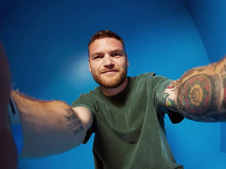 Mand tager selfie med udstrakte arme mod en blå væg. Han har tatoveringer på begge arme og bærer en grøn T-shirt.