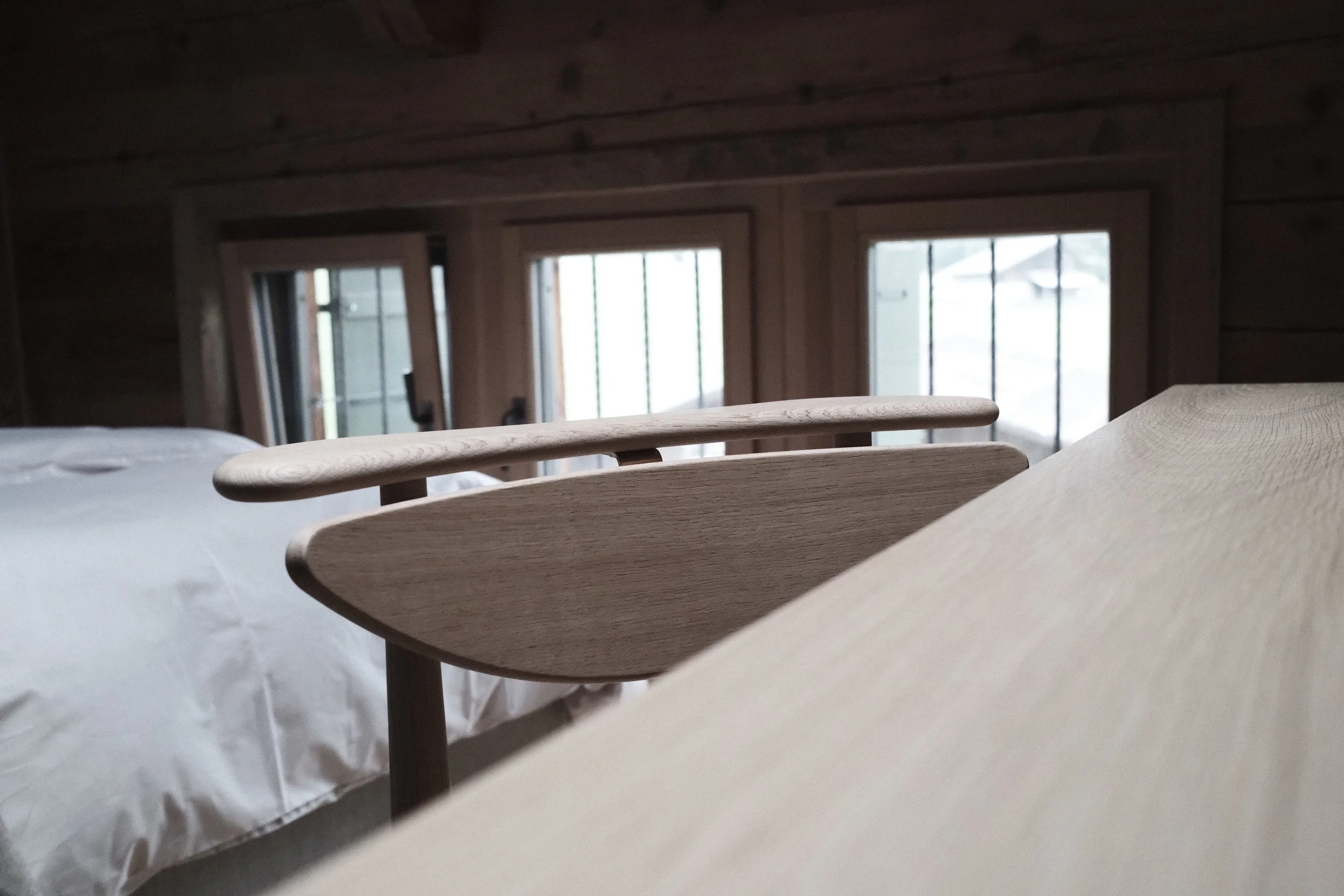 Close-up of a wooden bed frame with a white bedspread in a room with large windows and wooden walls.