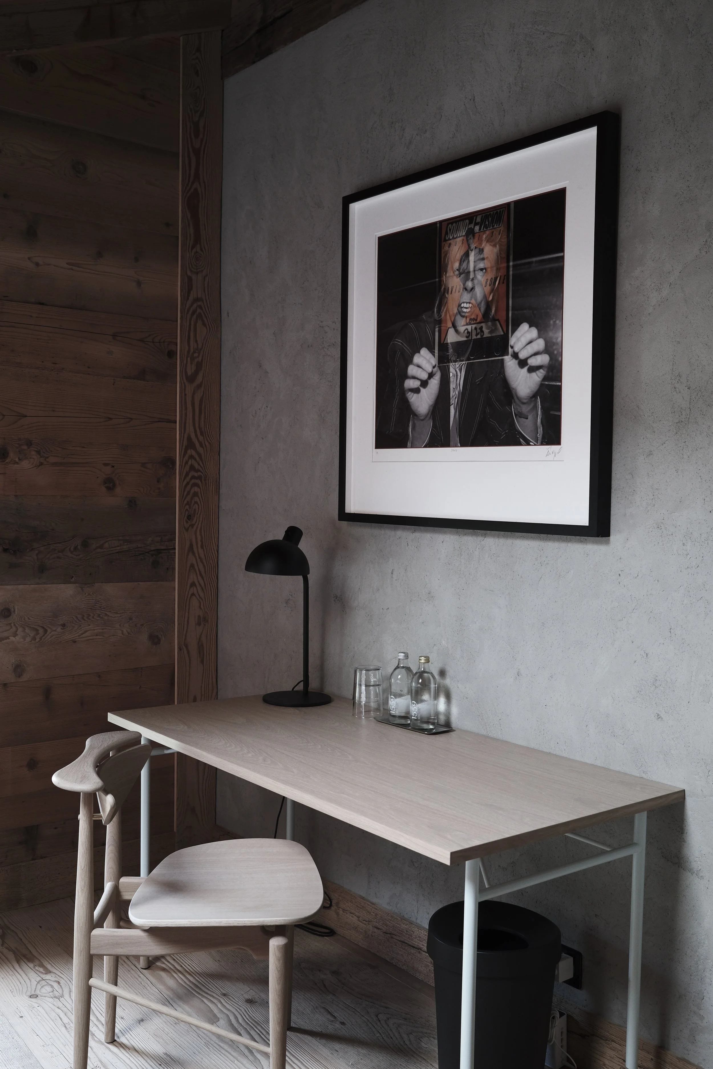 A modern dining area with a light wooden table, two matching chairs, a black desk lamp, and a framed black-and-white photo of a woman with a newspaper on her face hanging on a gray wall. There are water bottles and glasses on the table, and a black waste bin underneath.
