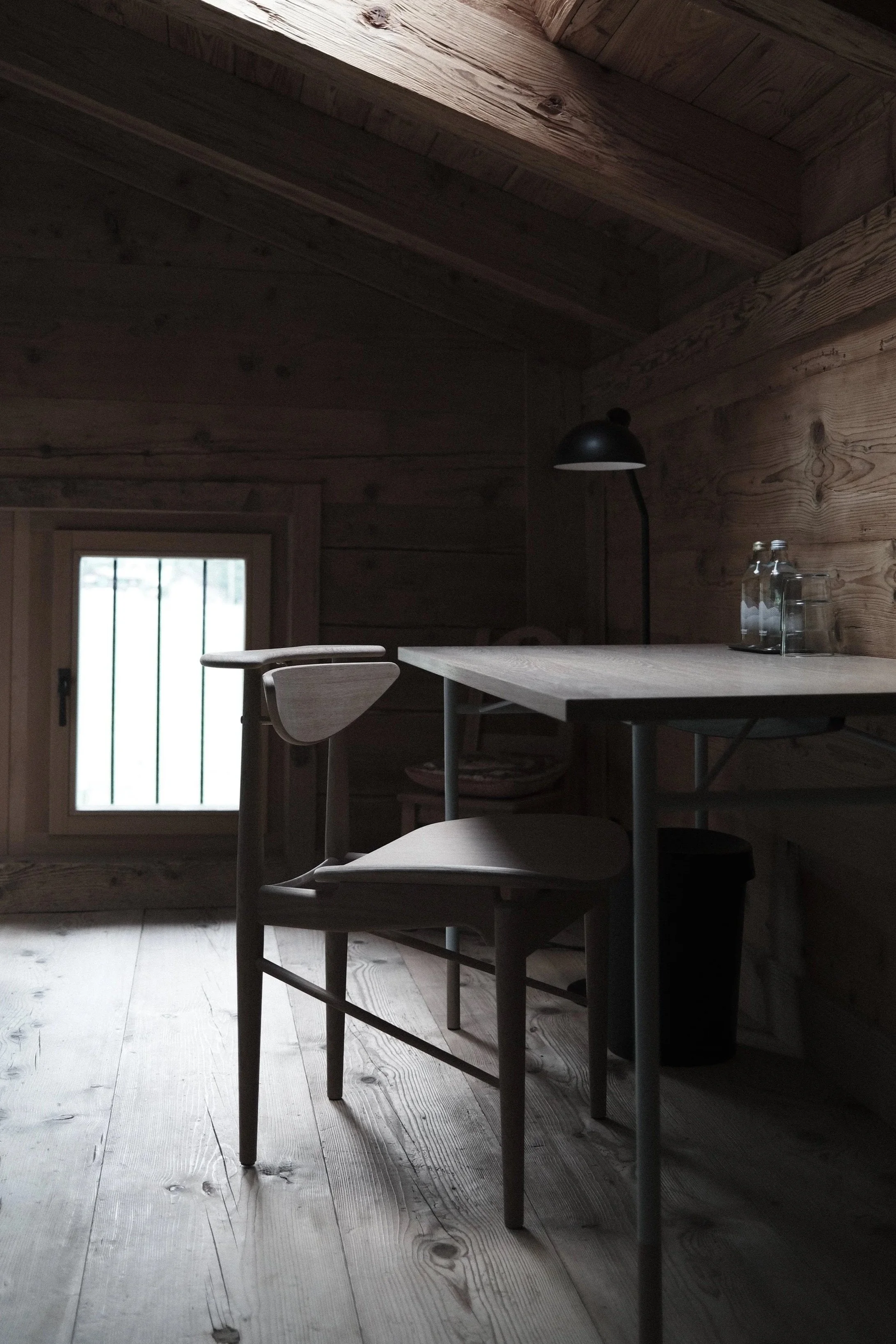 Interior of a wooden cabin with a table and chairs, a black desk lamp, and water bottles on the table. A window with vertical bars is visible in the background.