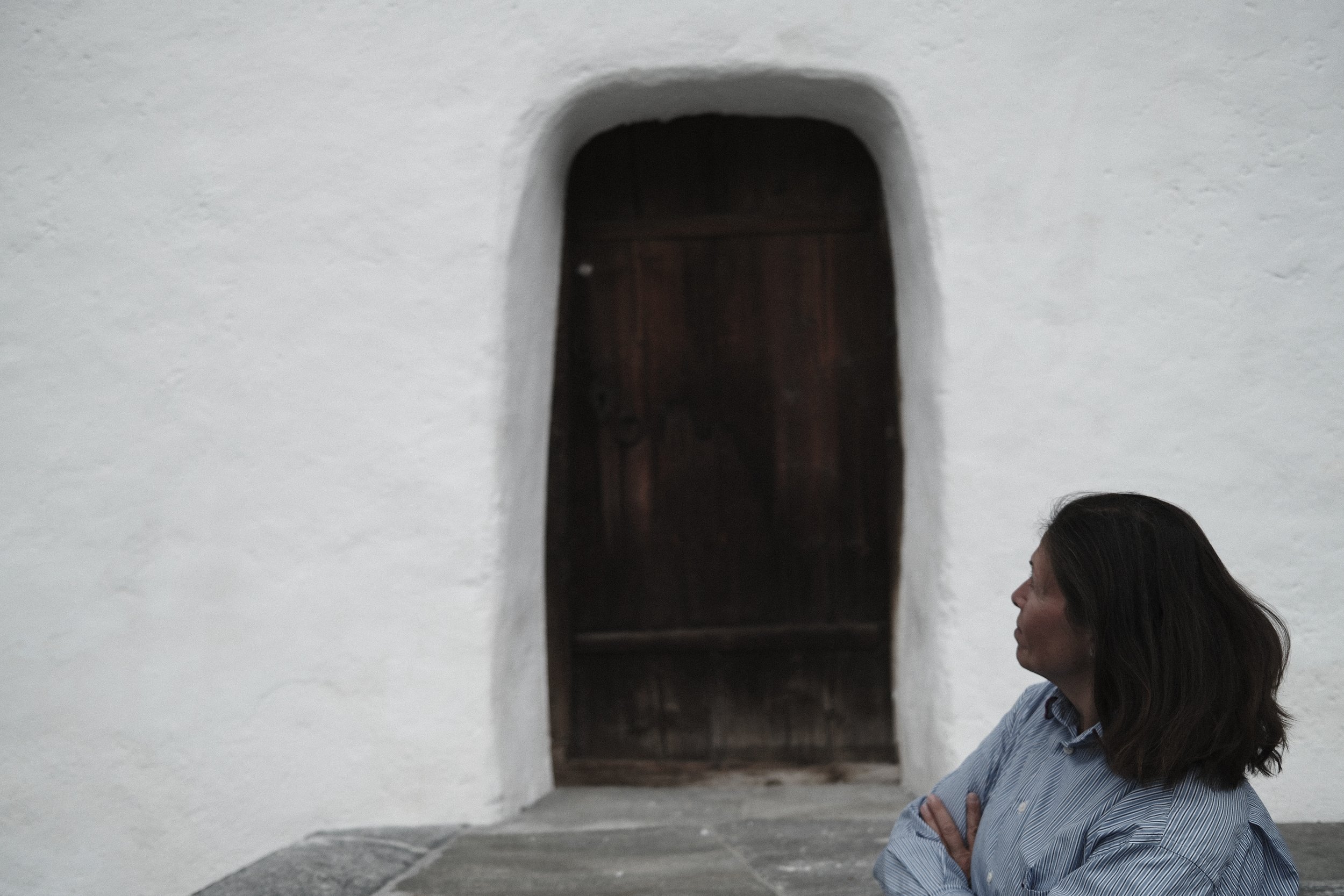 A woman with dark hair wearing a striped shirt stands with arms crossed in front of a white wall with an arched wooden door.