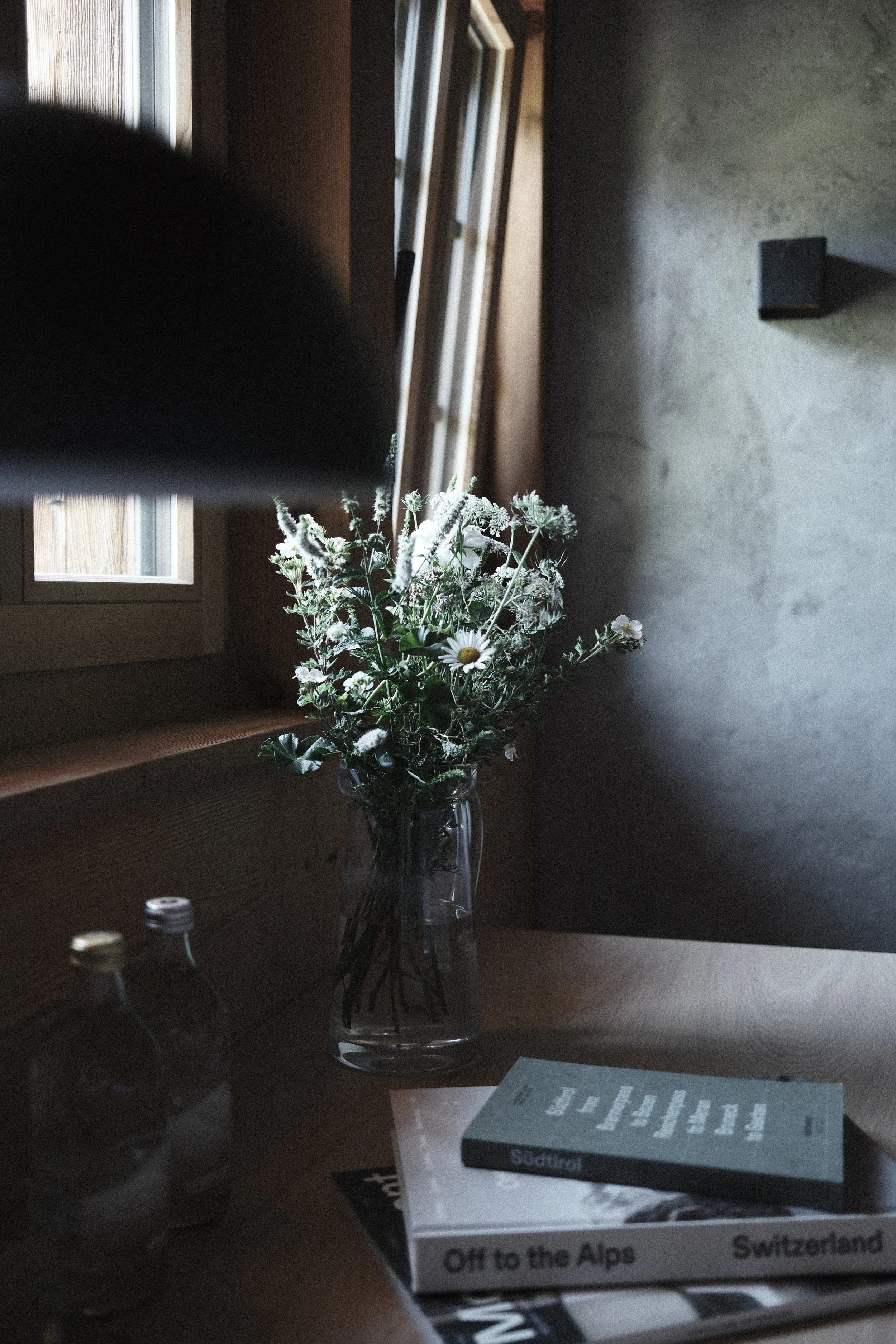 A clear glass vase with a bouquet of white daisies and greenery, placed on a wooden table near a window with wooden frames. There are a few small bottles in the foreground and books titled 'Off to the Alps' and 'Switzerland' on the table. The room has a simple, modern decor with a concrete wall.