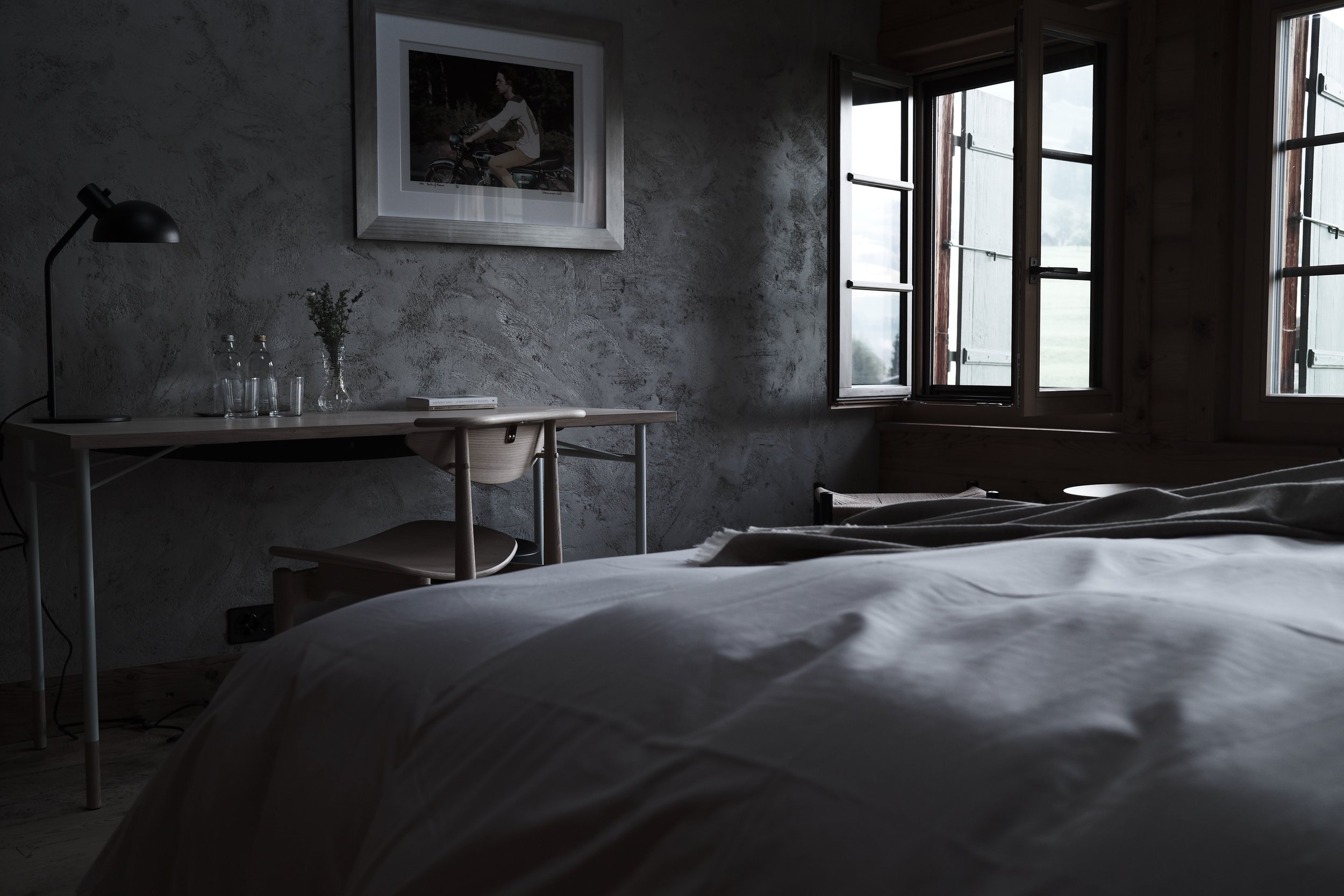 A cozy bedroom with a textured gray wall, a desk, a chair, a framed photo of a woman riding a bicycle, and an open window showing an outdoor view.