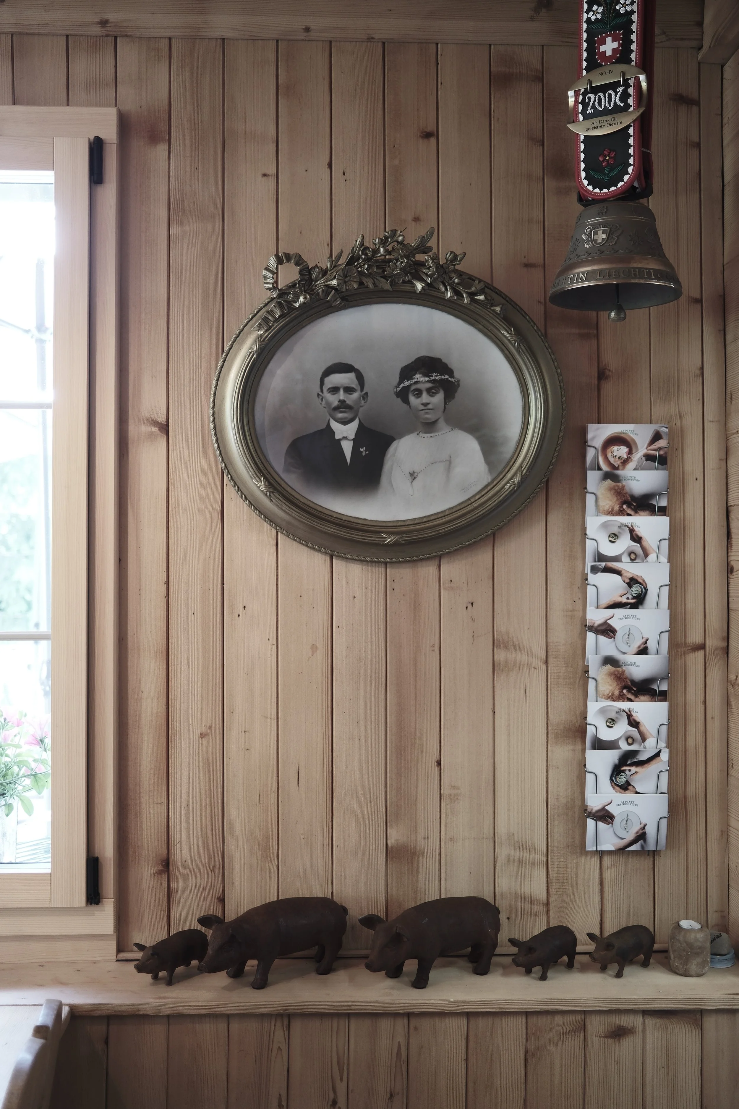 Wood-paneled wall with framed black-and-white portrait of a man and woman, a bell with Swiss Emblem, a brochure holder with various leaflets, three pig statues on shelf, and a window on the left side.