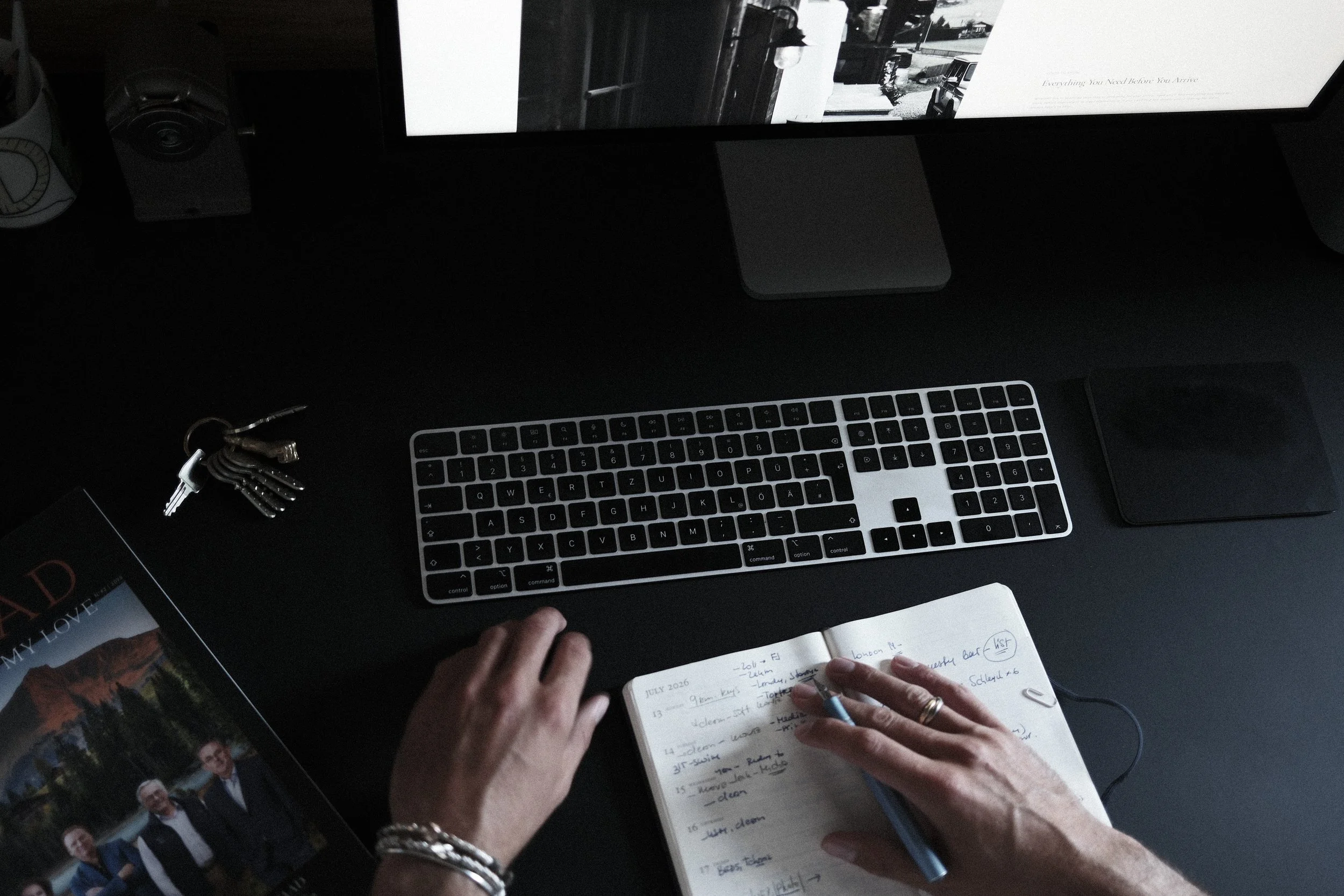 Person writing notes in a notebook in a workspace with a computer monitor, keyboard, and other items on a dark desk.