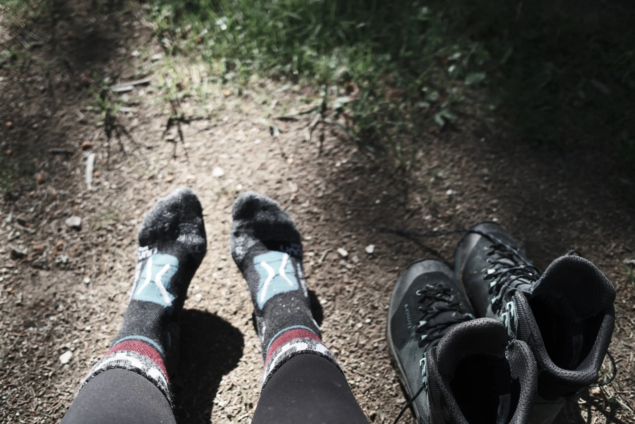 Photo of two pairs of feet wearing sneakers and socks, resting on dirt and small rocks in a forested area.
