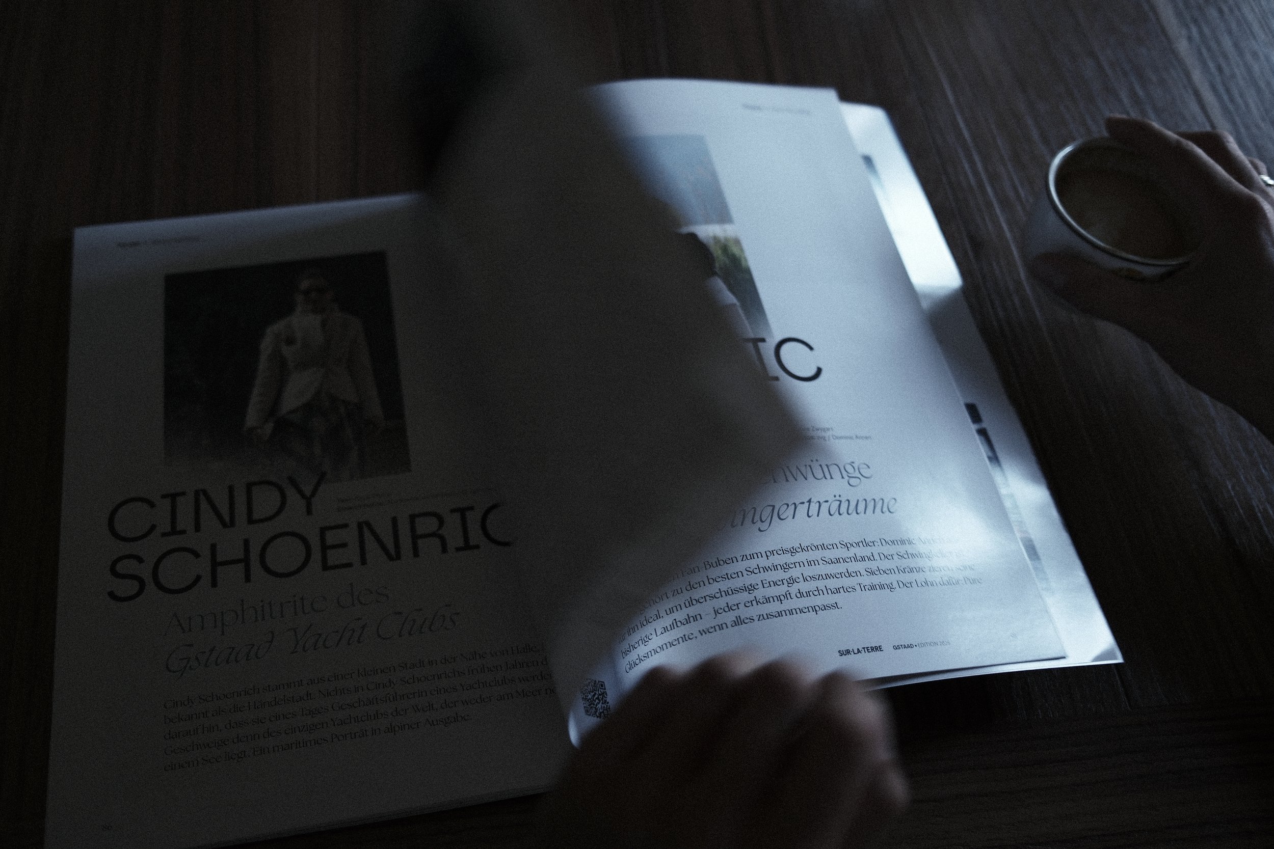 Open magazine or booklet with German text, photo of a man in a suit on the cover, and a person's hand holding a small black object, possibly a cup, on a dark wooden surface.