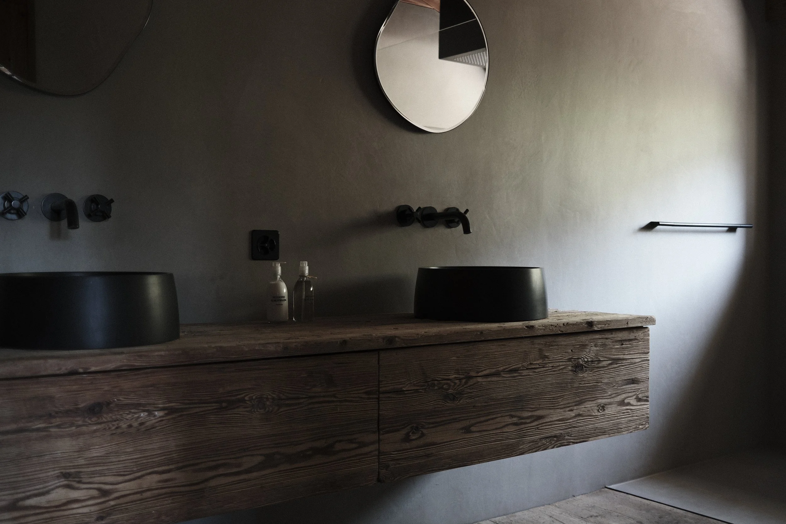 A minimalist bathroom vanity with a wooden cabinet, black vessel sinks, and black wall-mounted fixtures, including faucet and mirror, against a plain gray wall.