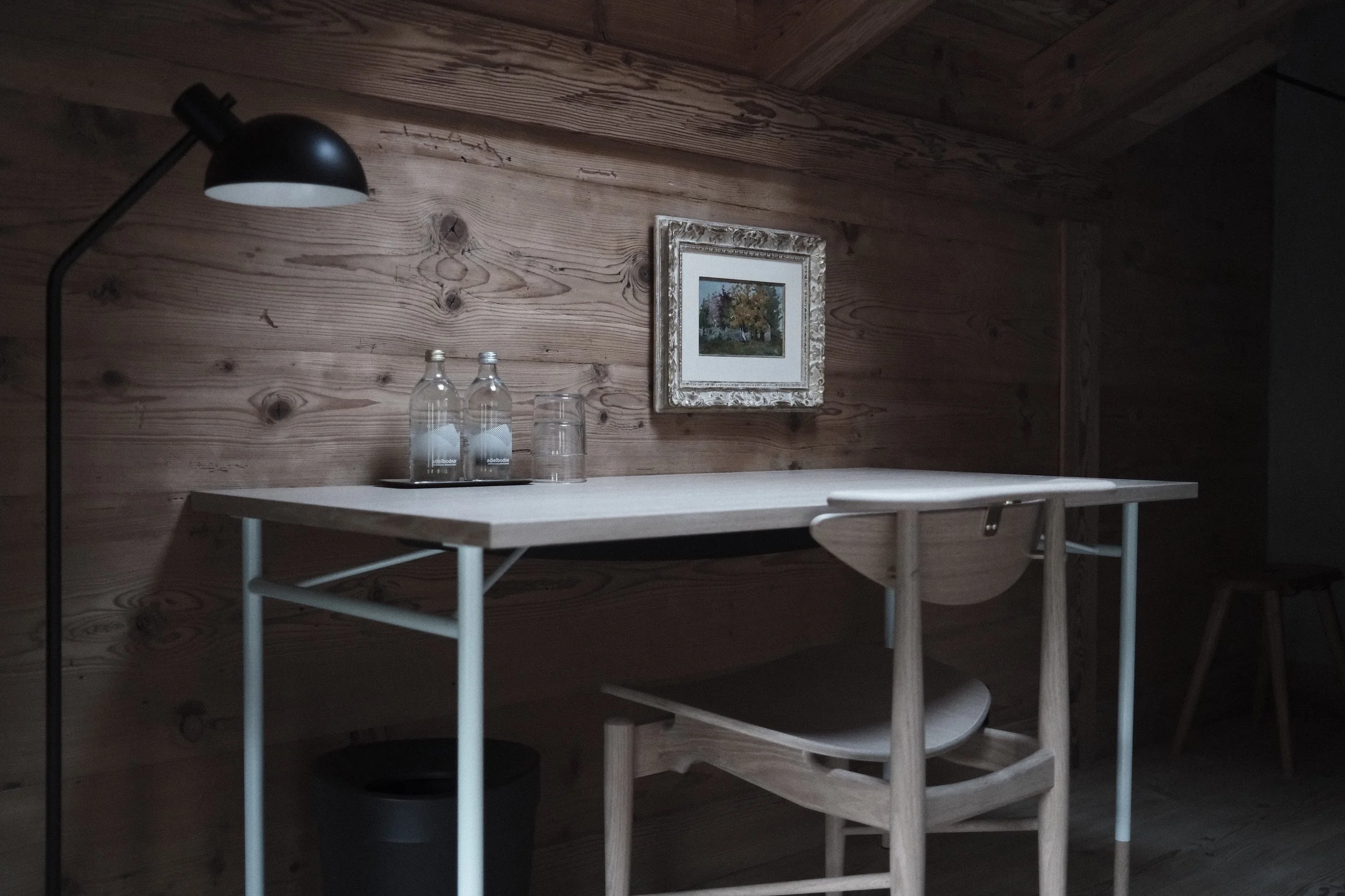 A wooden wall with a framed painting of trees, a black desk lamp, three glass bottles, and a glass on a white table.