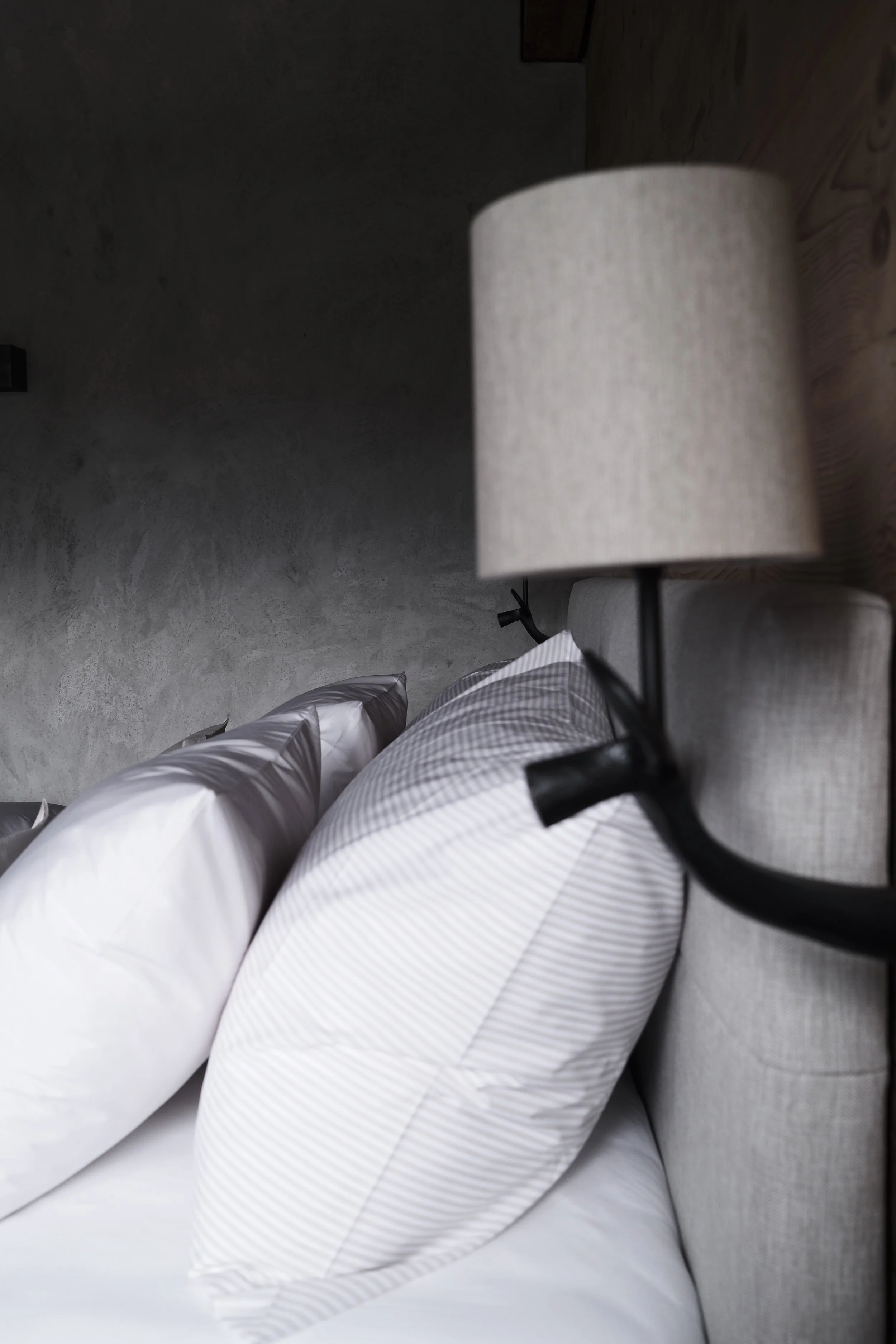 Close-up of a bed with white and gray striped bedding, a beige lampshade on a black lamp, and a modern gray textured wall in the background.