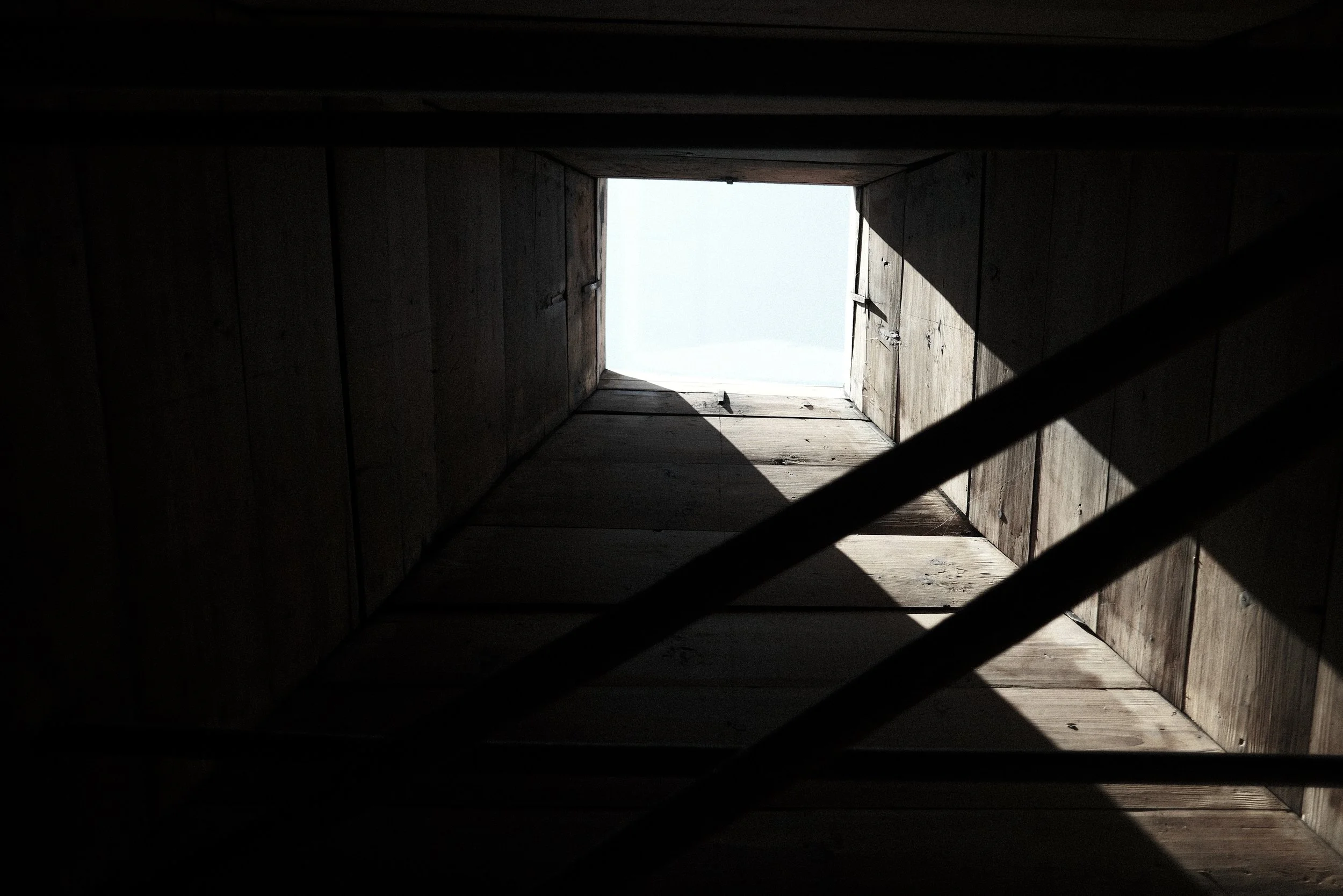 Looking up from inside a wooden attic or loft through an open square hatch in the ceiling, showing the sky outside.