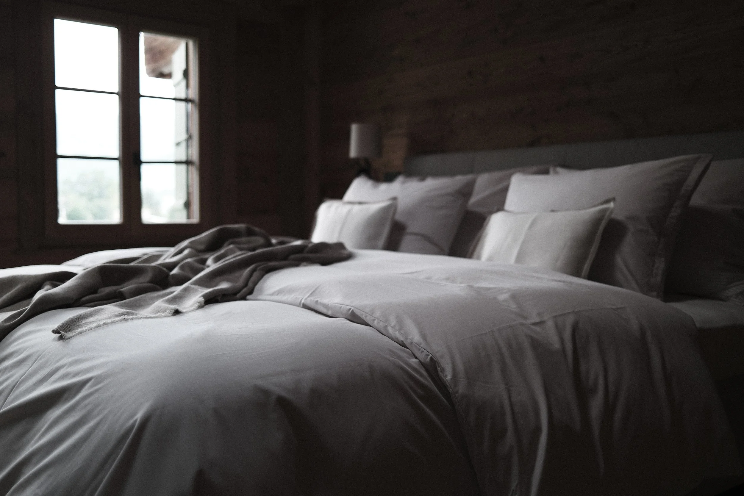 A bedroom with a wooden wall, bed with white sheets, pillows, and a gray blanket, and a window letting in natural light.