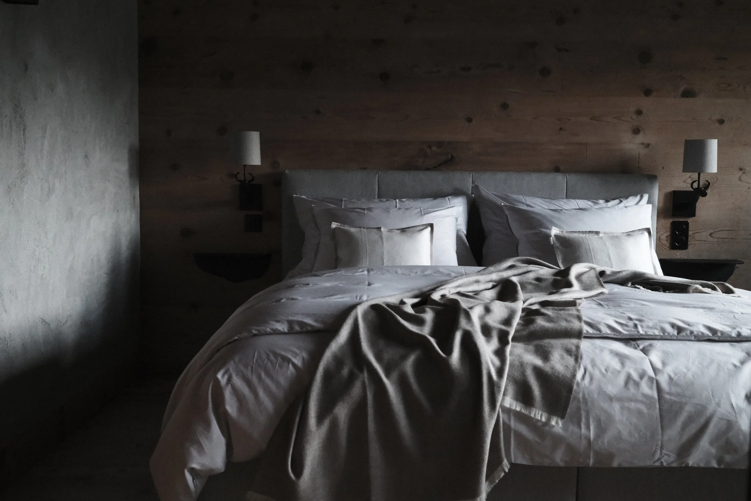 A bed with white linens and pillows, set against a wooden headboard and adjacent wall, with two wall-mounted lamps on either side in a room with dim lighting.