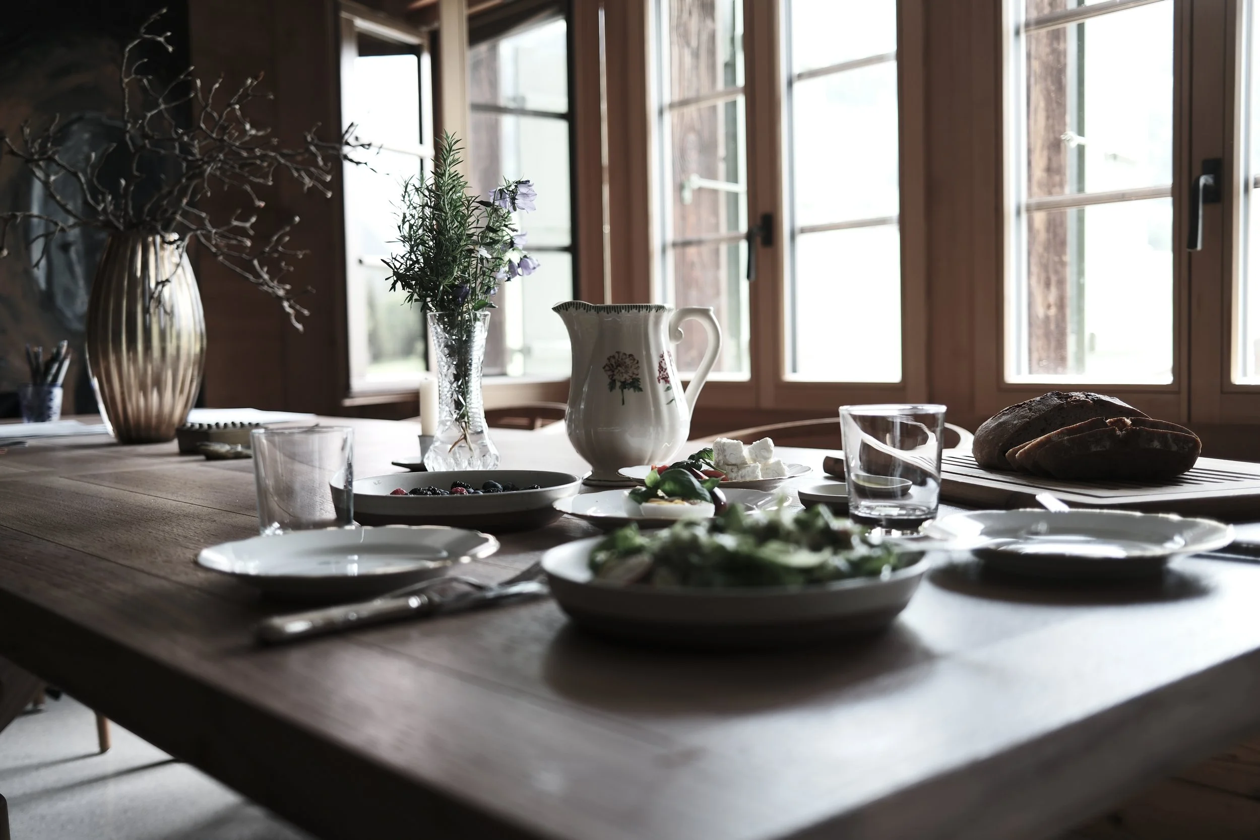A wooden dining table set with plates, glasses, a loaf of bread, a salad, and a pitcher, in front of large windows in a cozy, rustic kitchen or dining area with natural light.