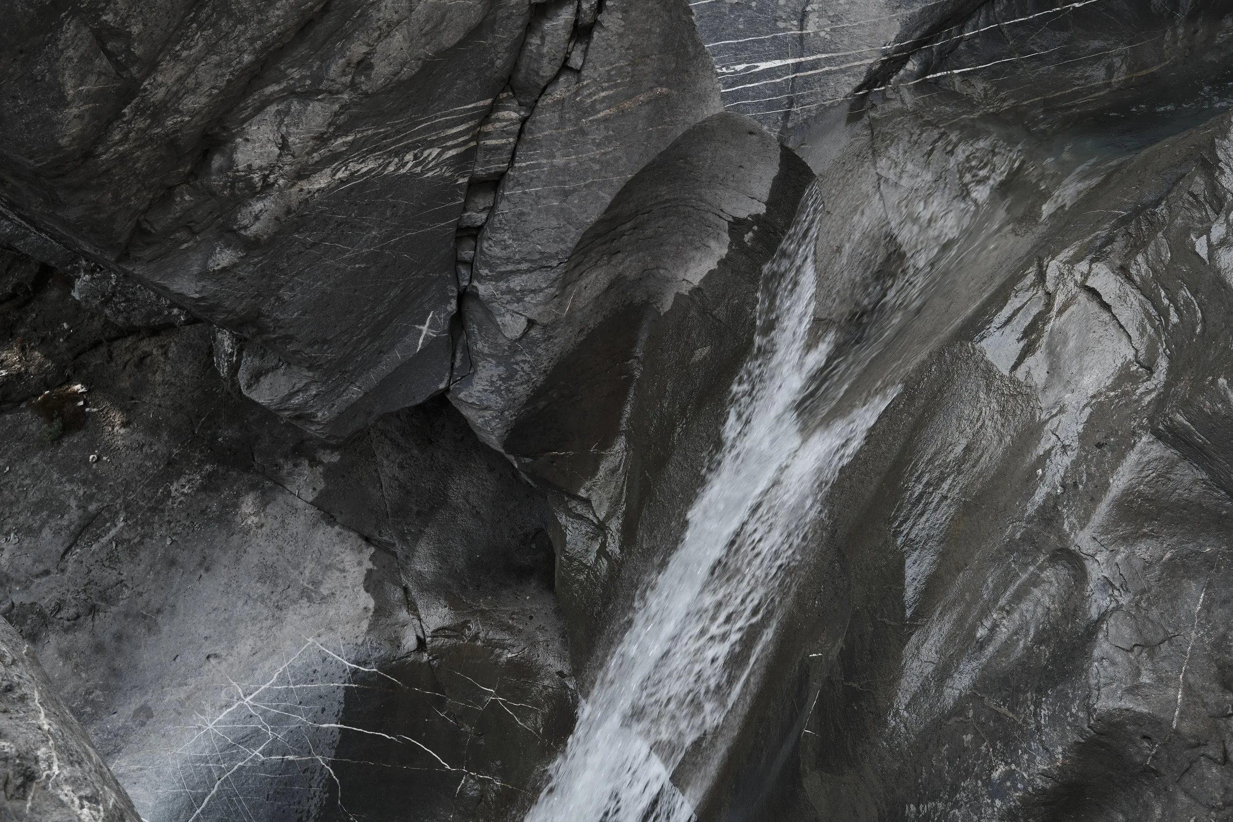 Close-up of dark gray rocks with a small stream of water flowing over them, revealing layered and cracked surfaces.