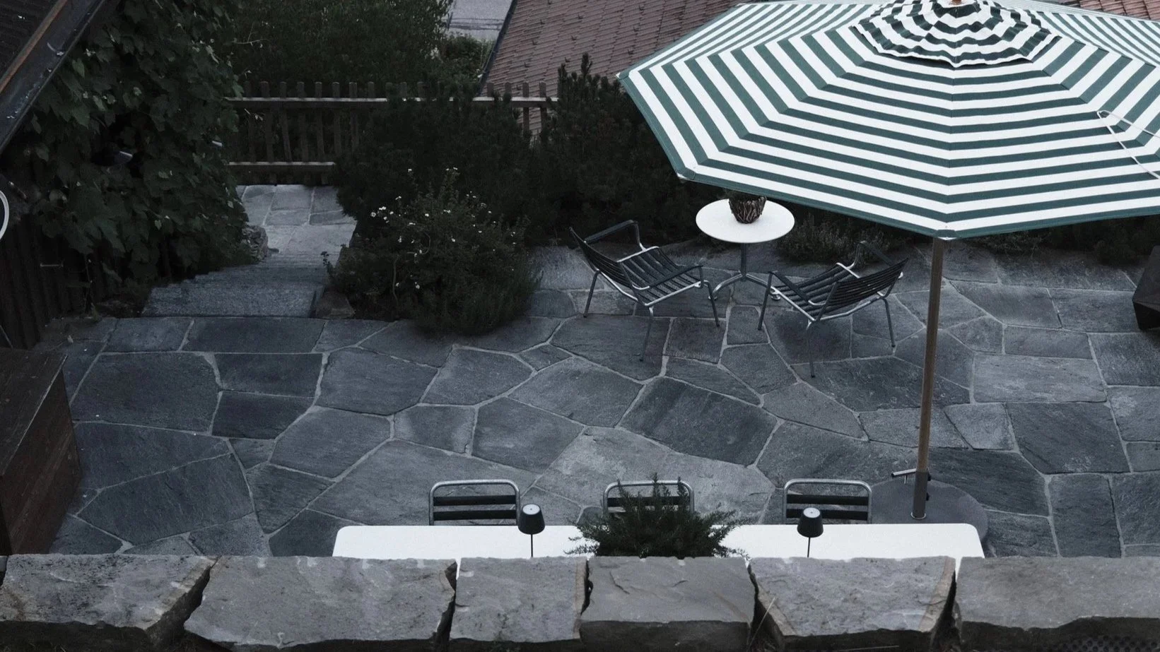 A stone patio with a white table, black chairs, a large green and white striped umbrella, and some greenery in a backyard.