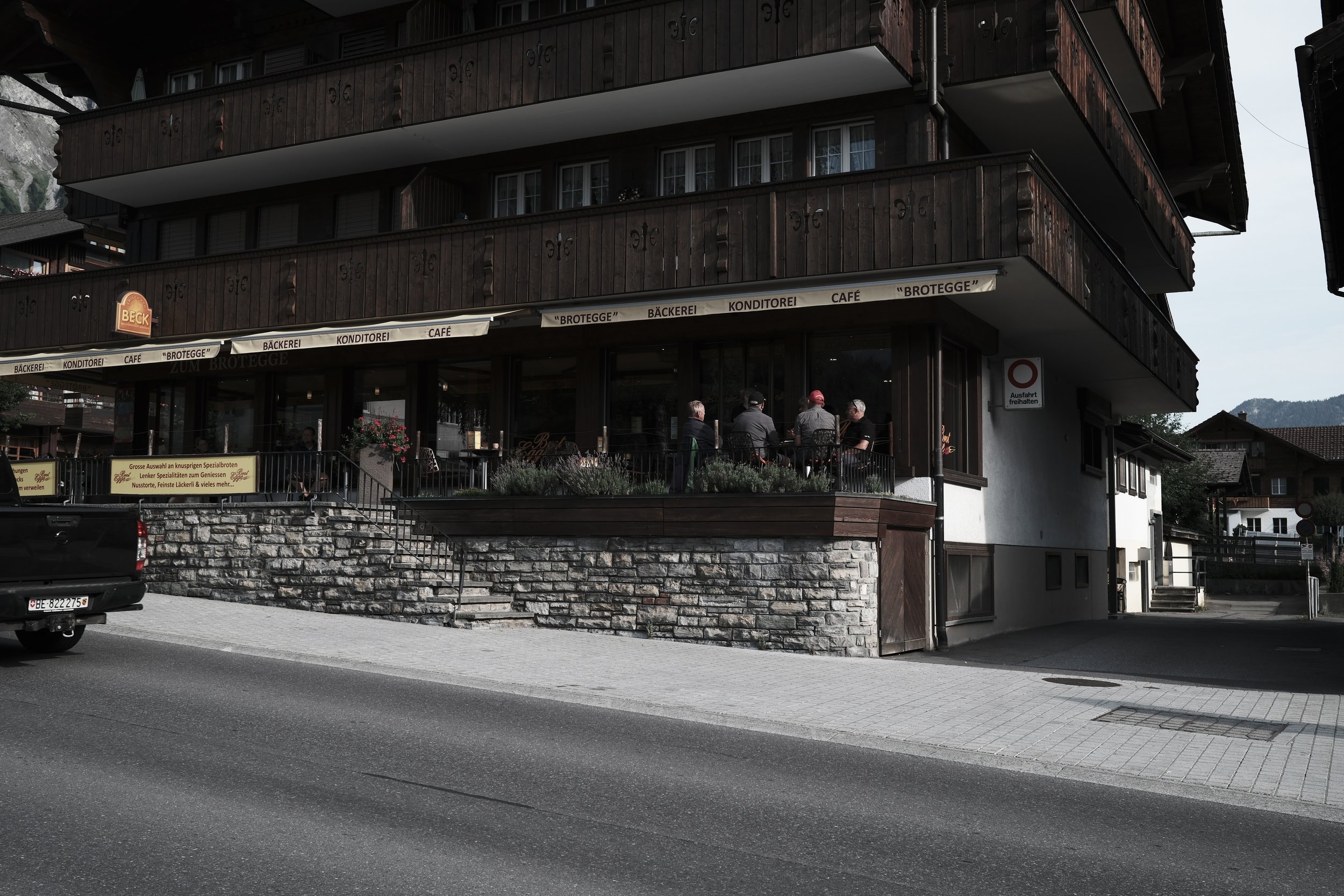 A multi-story building with wooden balconies, a cafe with outdoor seating, and a stone foundation. Several people are sitting at the cafe, and there is a sign with German words.