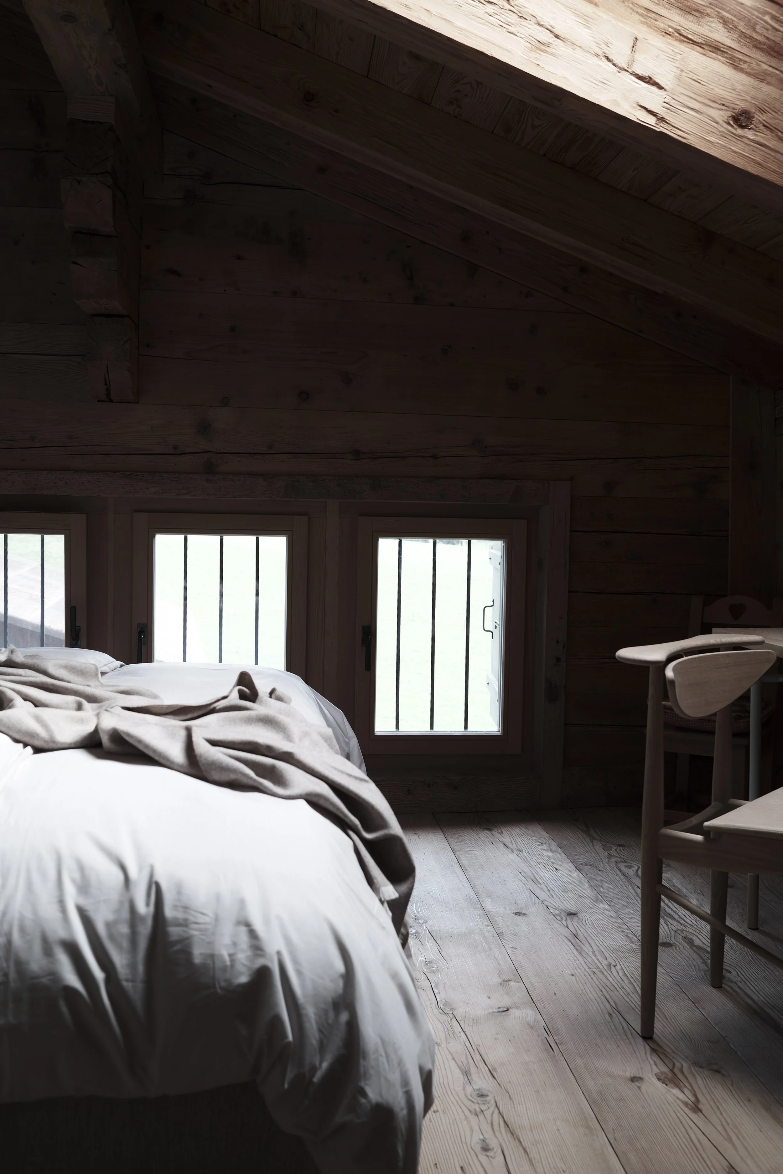 A cozy room featuring a sloped wooden ceiling with natural wood planks, three windows with black bars, a bed with white bedding and a beige blanket, and a light wooden chair.