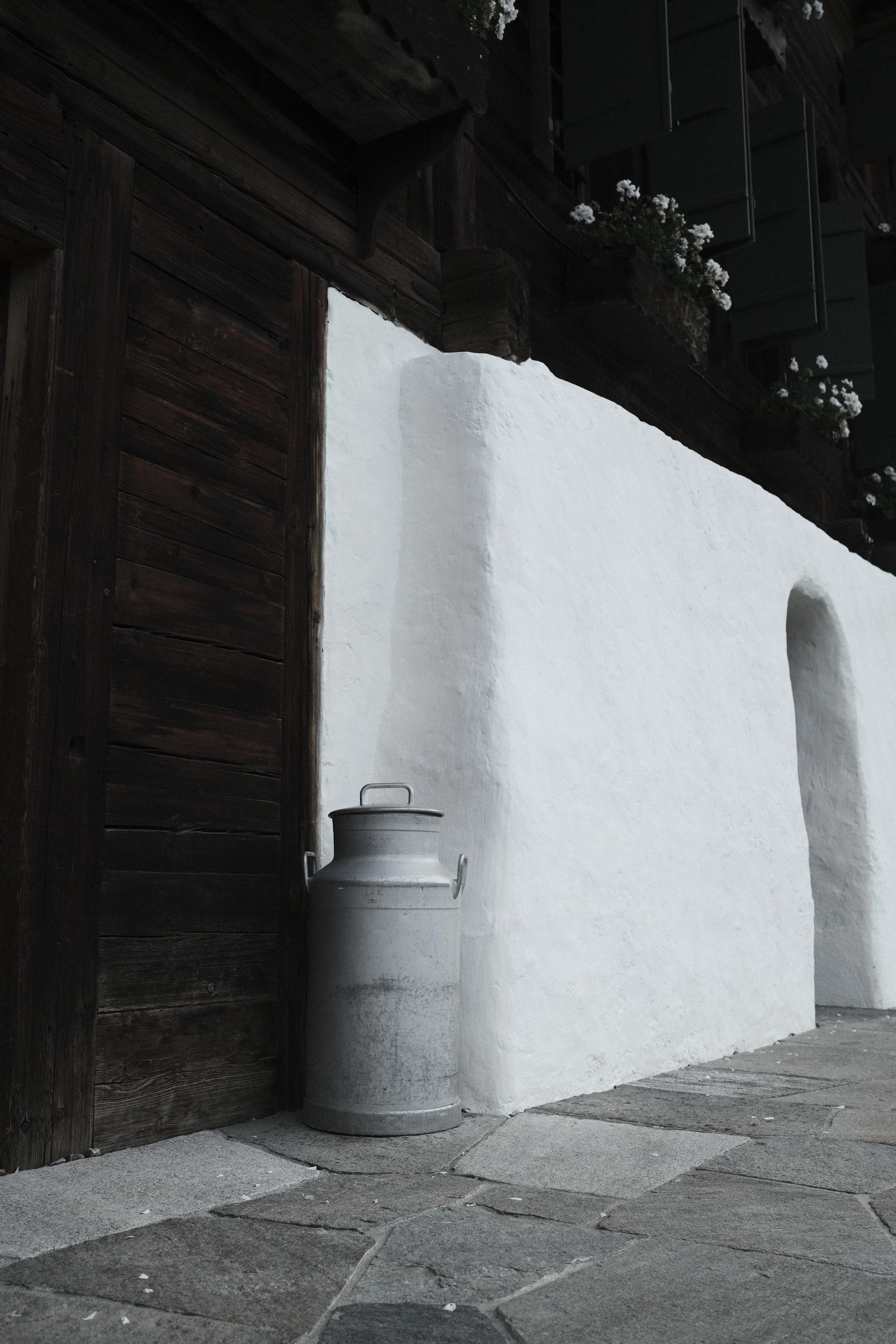 A rustic doorway with dark wooden shutters and a white wall, featuring a metal milk can and flower boxes with white flowers.
