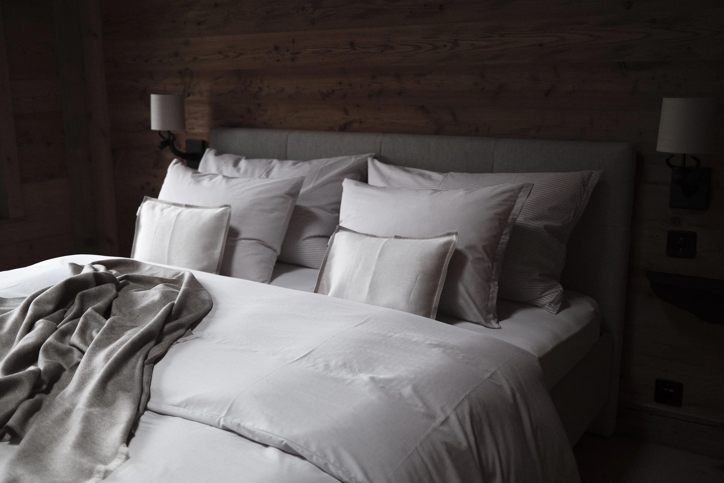 A neatly made bed with white and beige pillows, a gray headboard, and a tan blanket, set against a wooden-paneled wall in a bedroom.