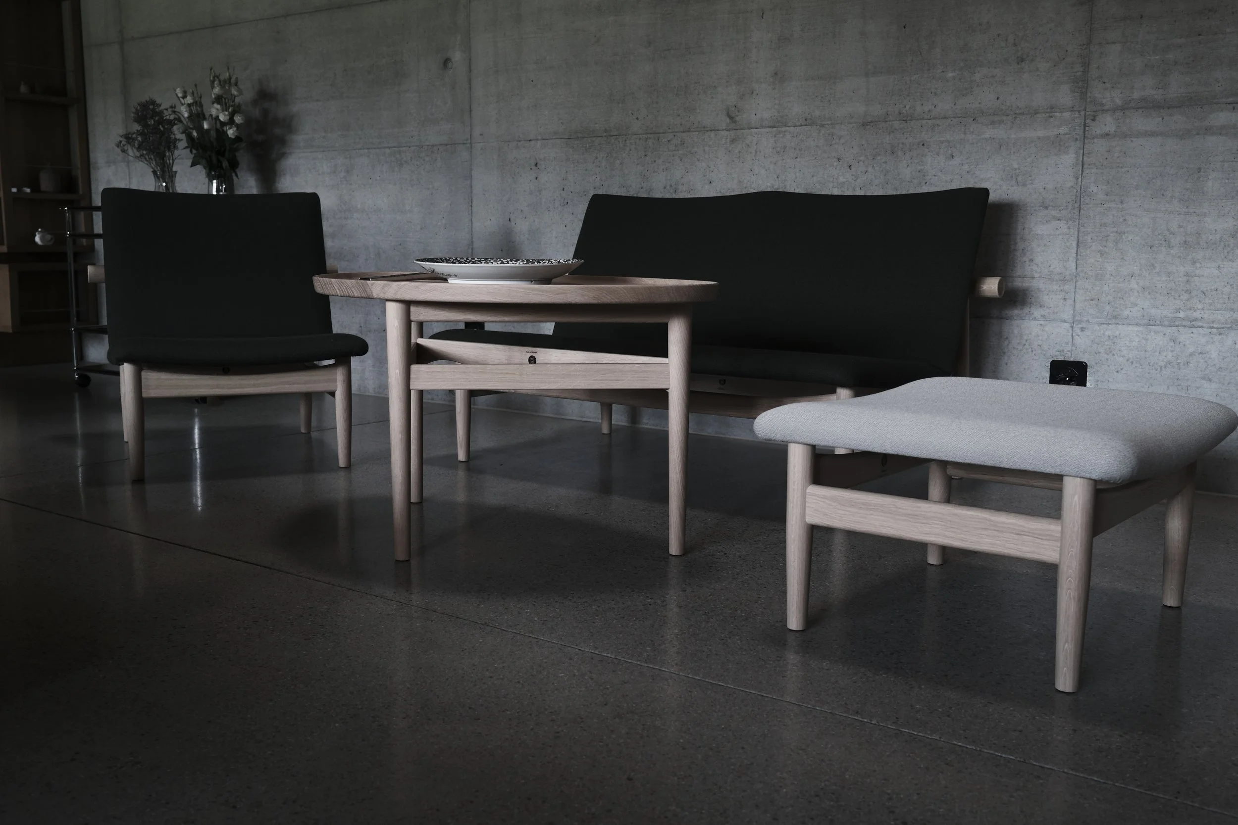 Minimalist living room with black armchair, light wood coffee table with a decorative plate, gray upholstered bench, concrete wall, and a vase with flowers in the background.