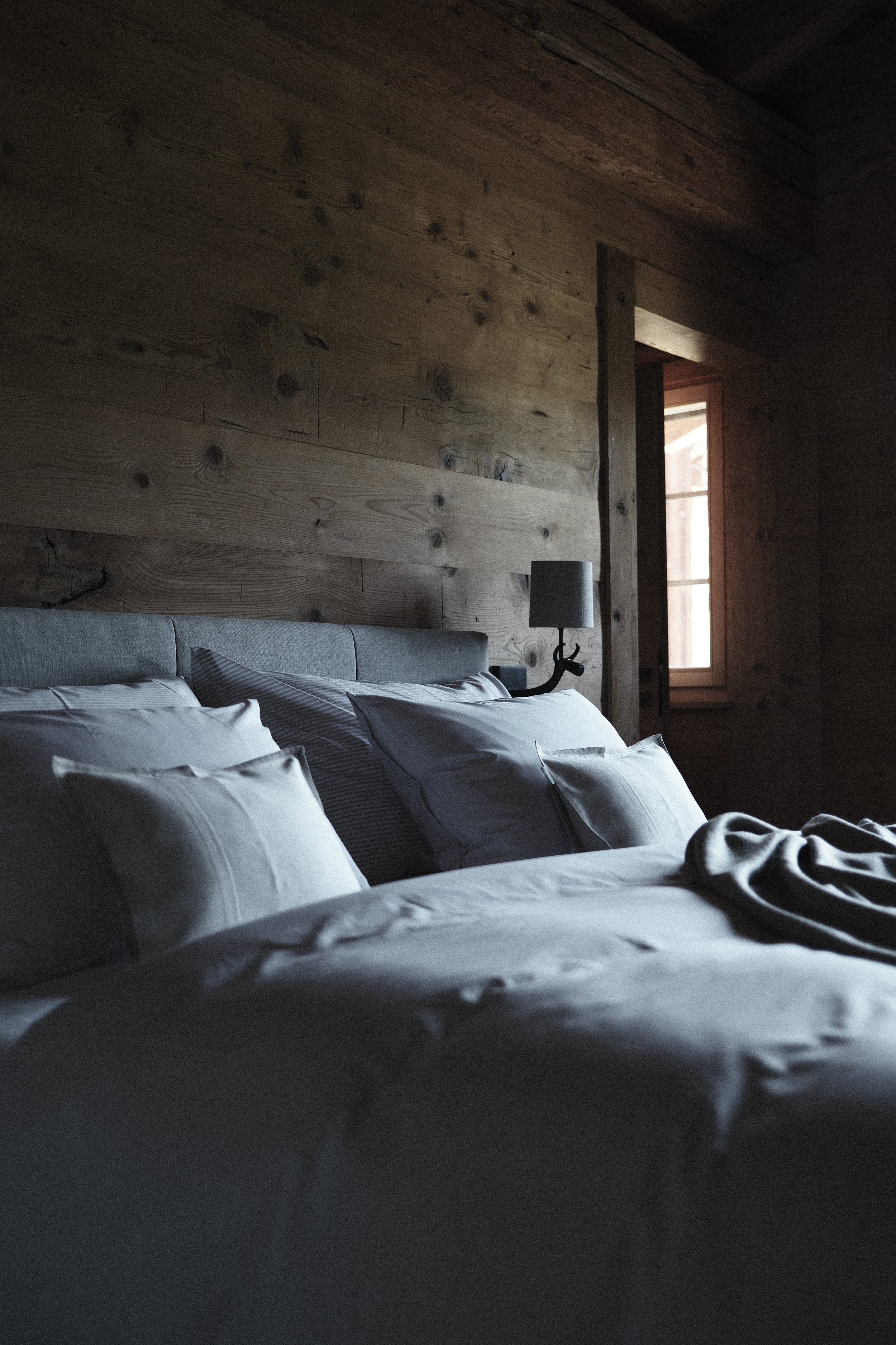 A bedroom with a wooden wall, a bed with white and gray bedding, pillows, a nightstand with a gray lamp, and a window letting in natural light.