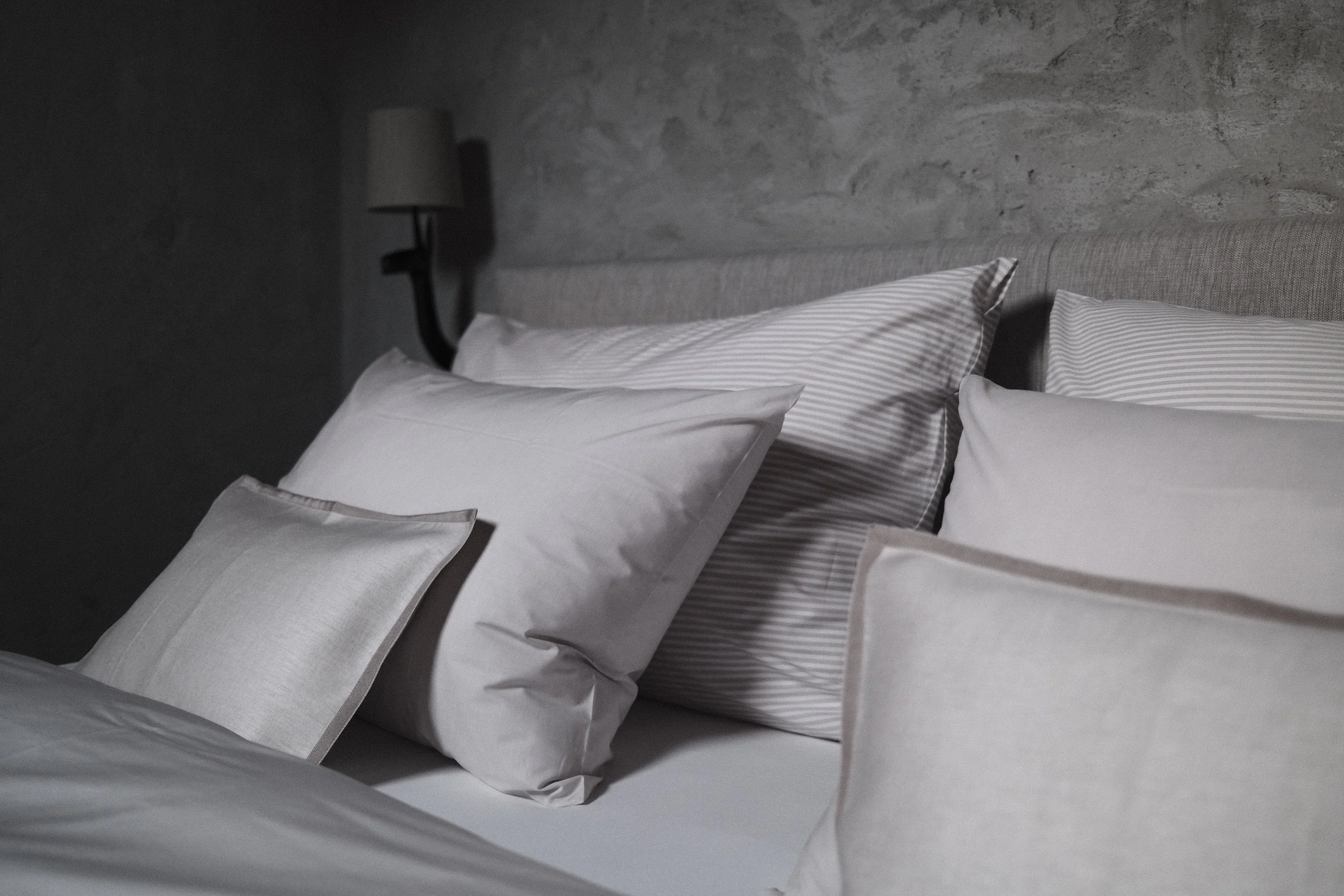 A neatly made bed with white sheets, striped blankets, and several pillows, set against a textured stone wall with a wall-mounted lamp beside it.