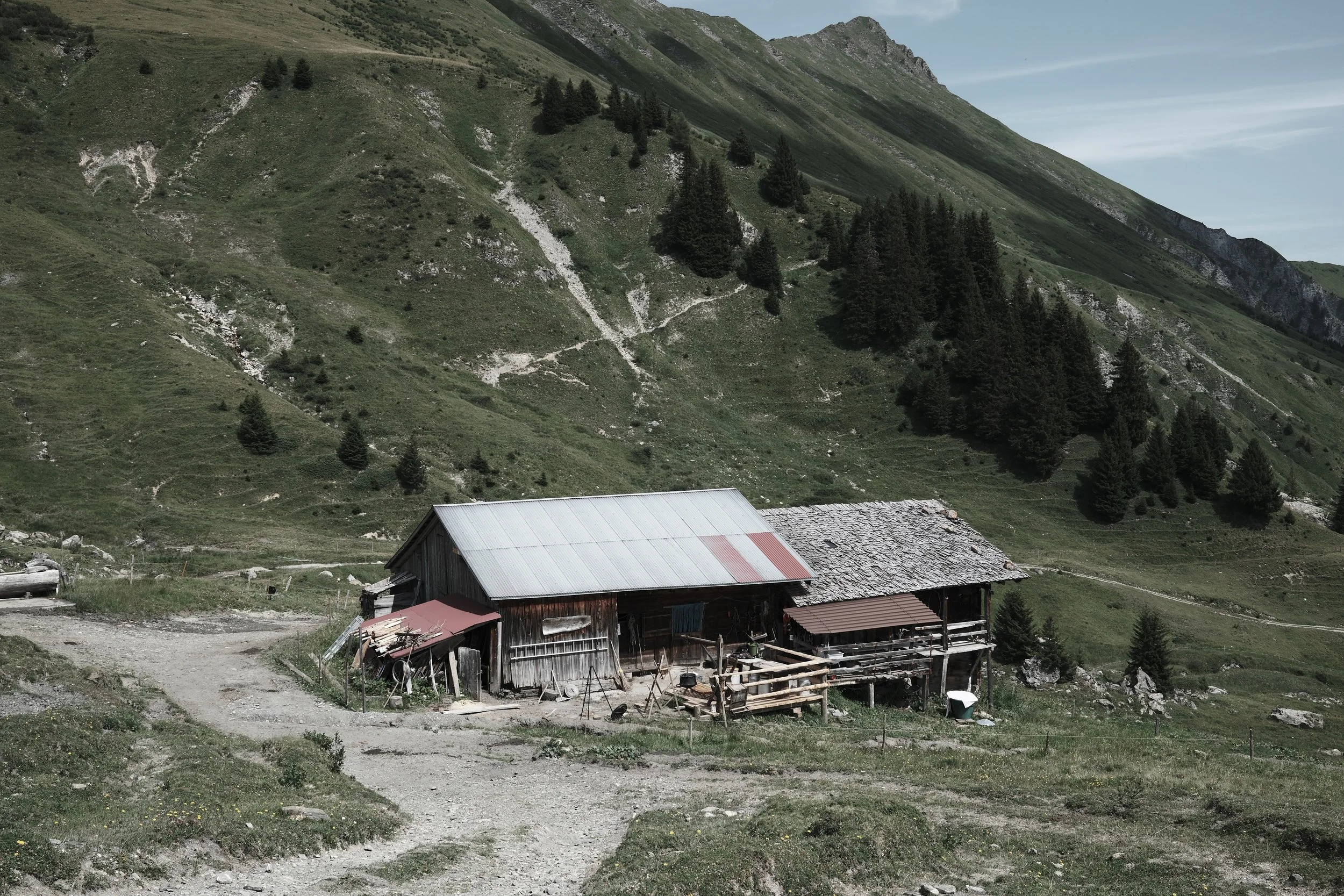 A small rustic wooden house with a metal roof, situated in a mountainous area with lush green slopes and scattered trees, under a partly cloudy sky.
