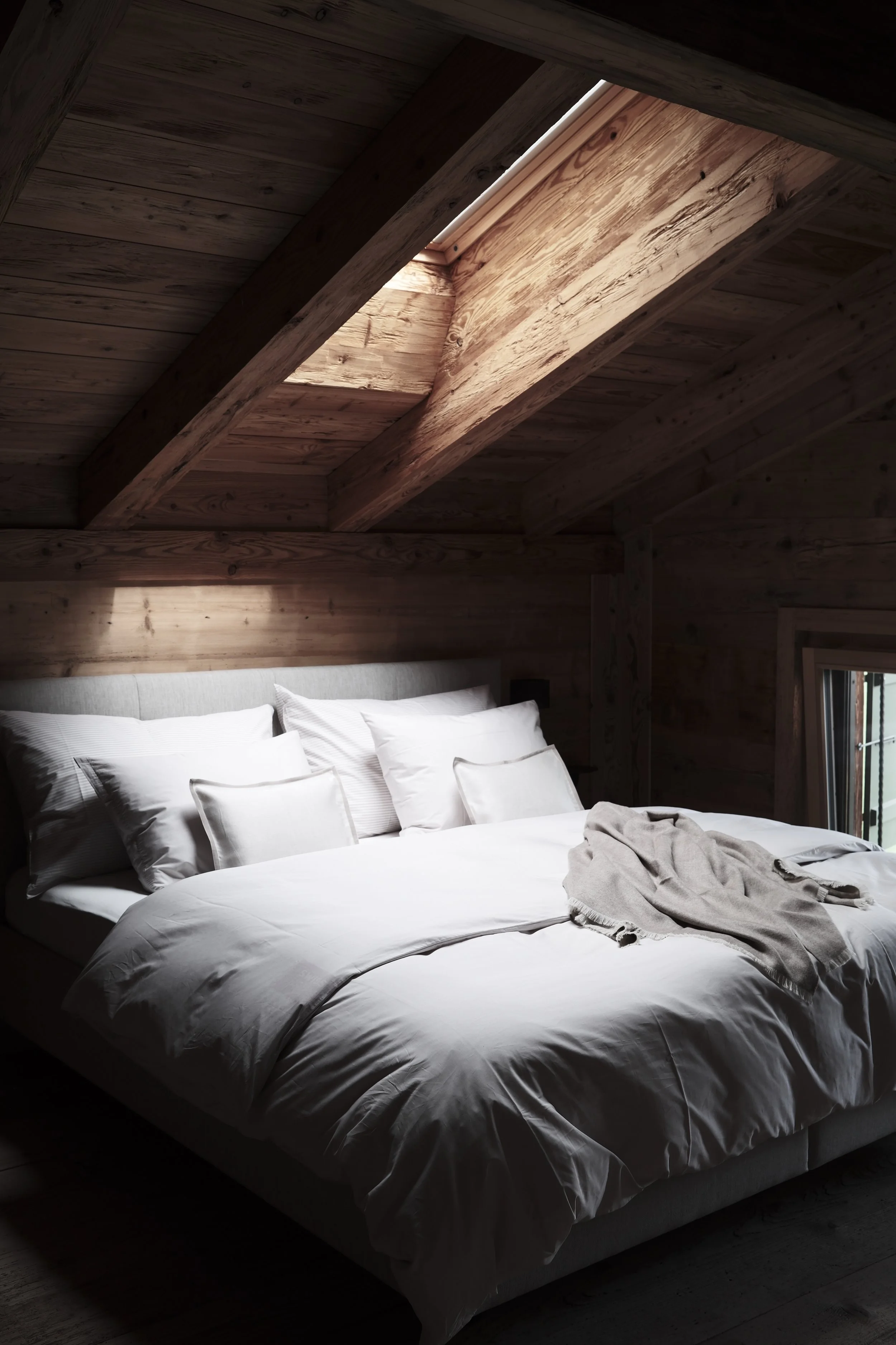 A cozy bedroom with a bed made up with white sheets and pillows, a light gray blanket on the bed, set inside a wood-paneled room with a slanted wooden ceiling and a small window.