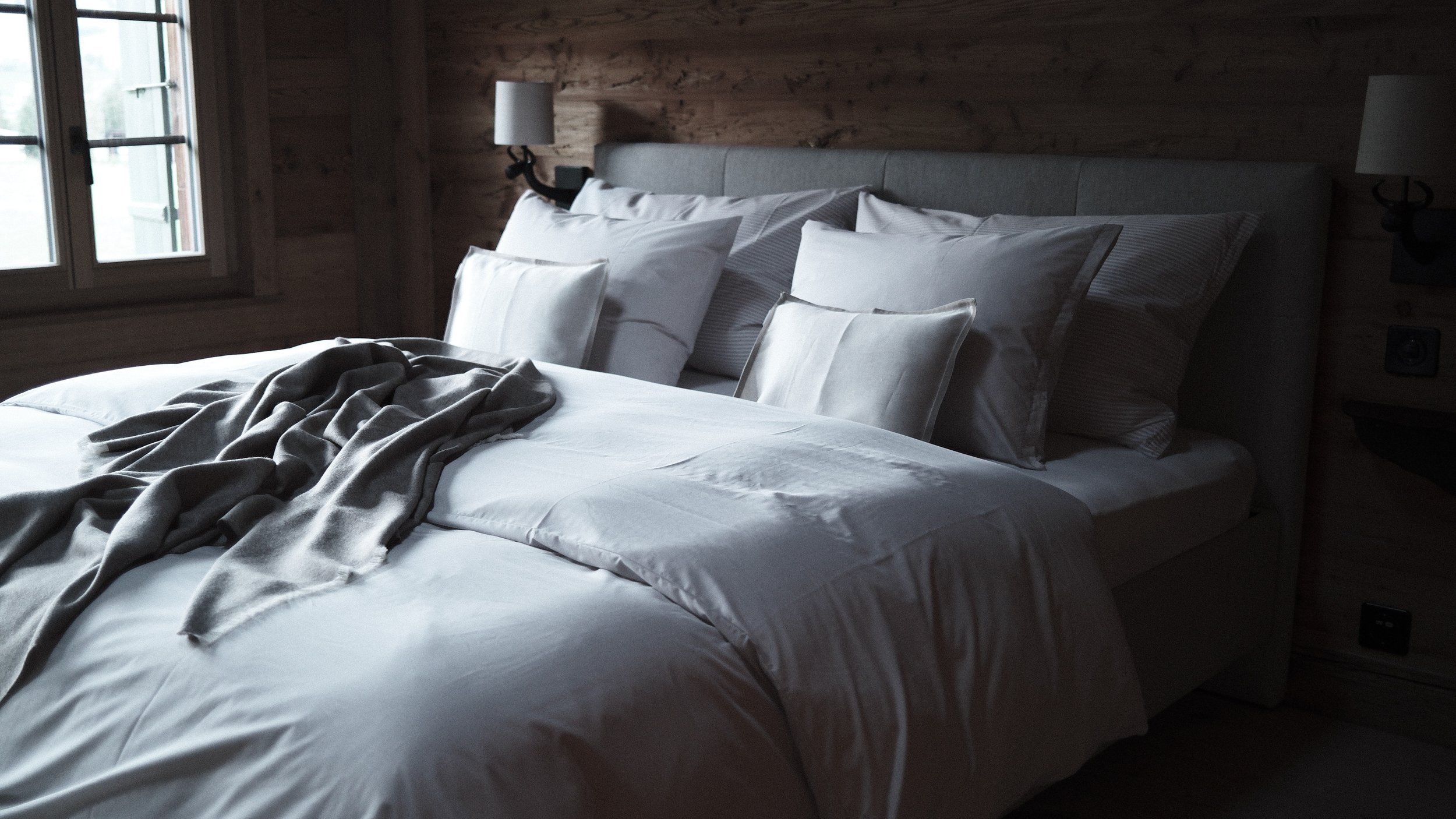 A neatly made bed with white sheets and multiple pillows in a room with wooden walls and a window.