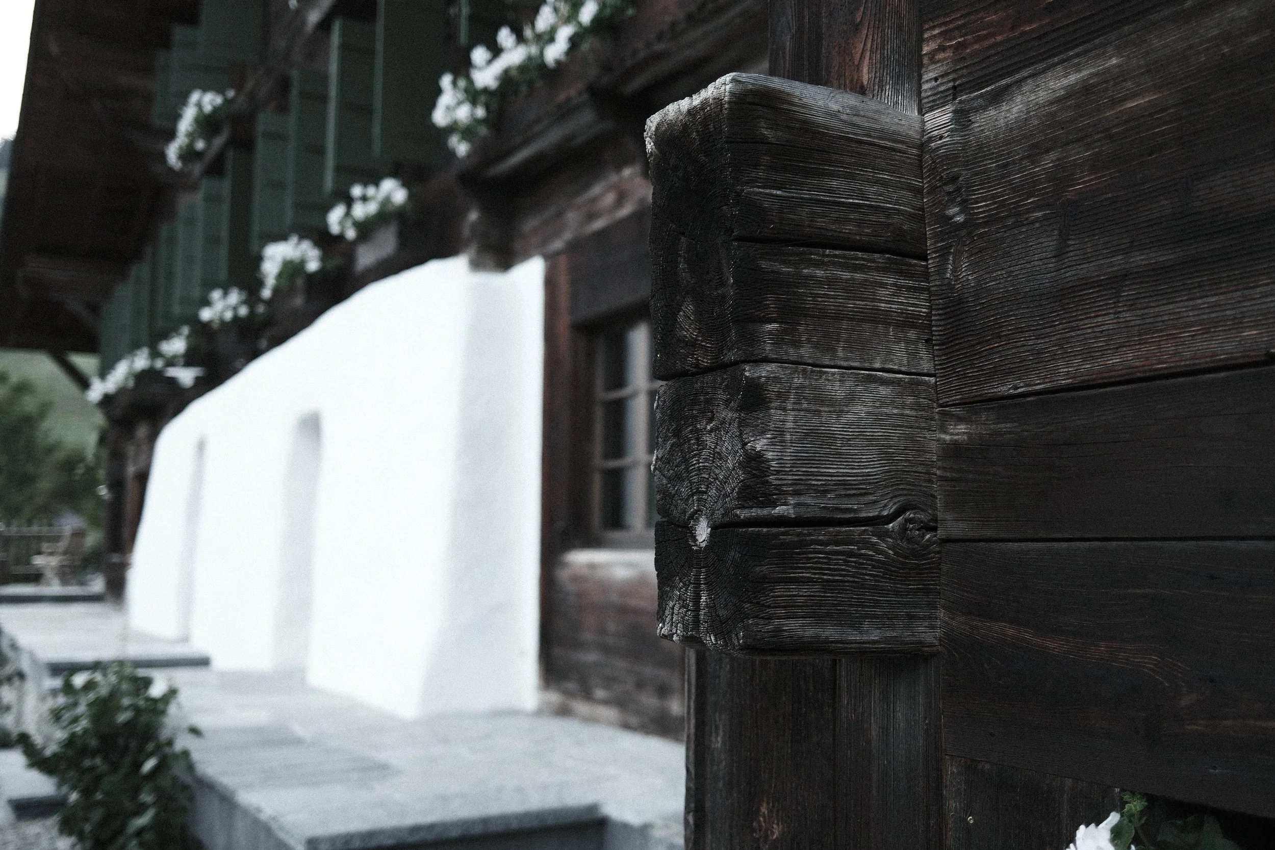 Close-up of weathered dark wooden beam of a house with exterior wall in background, outdoor scene with plants and a car.