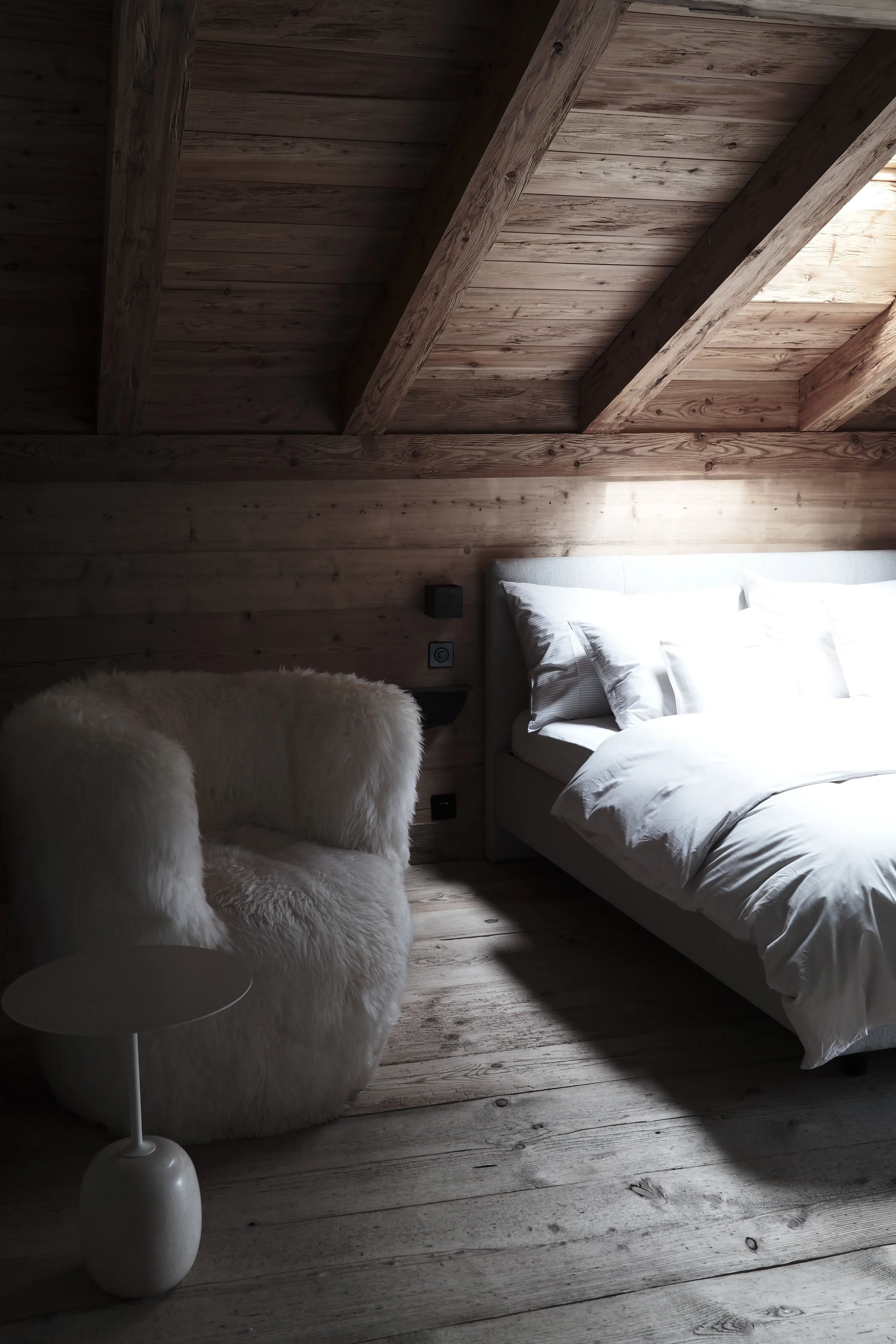 A cozy bedroom with wooden walls and ceiling. There is a bed with white bedding and pillows. Next to the bed, there is a fluffy white armchair with a small round white side table and a modern white lamp. Sunlight filters into the room, highlighting the wood textures.
