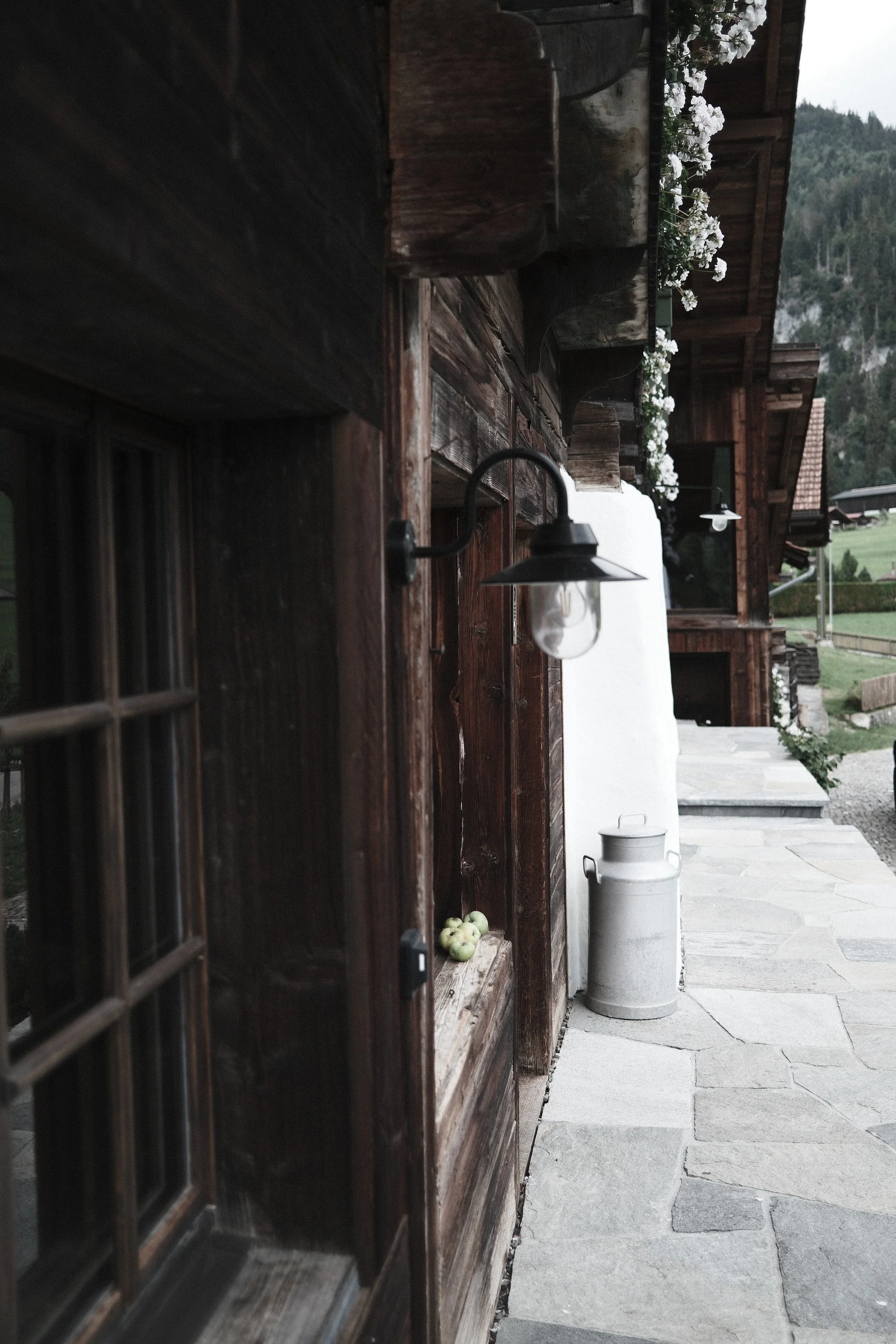A rustic wooden exterior wall with a window, a vintage-style outdoor lantern, a white container, and some apples, with a mountain landscape in the background.