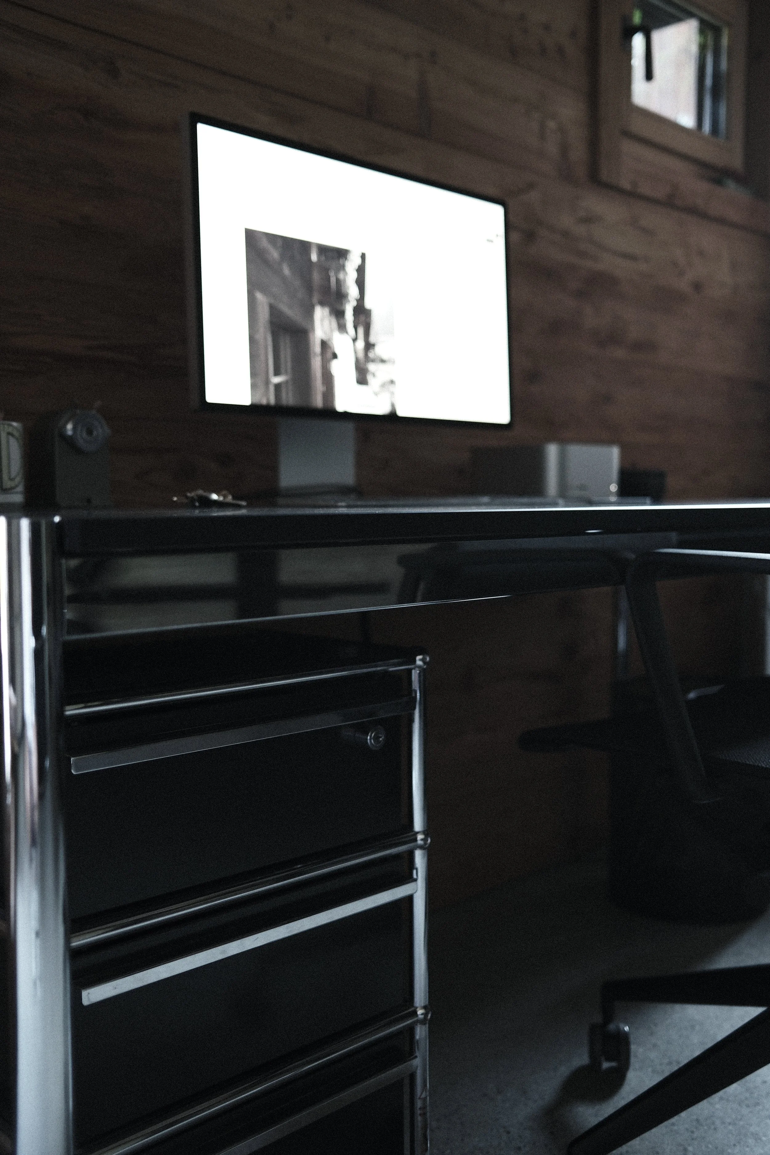 Black rolling tool cabinet with multiple drawers in front of a wooden wall, with a computer monitor on a desk to the right and small framed pictures above.