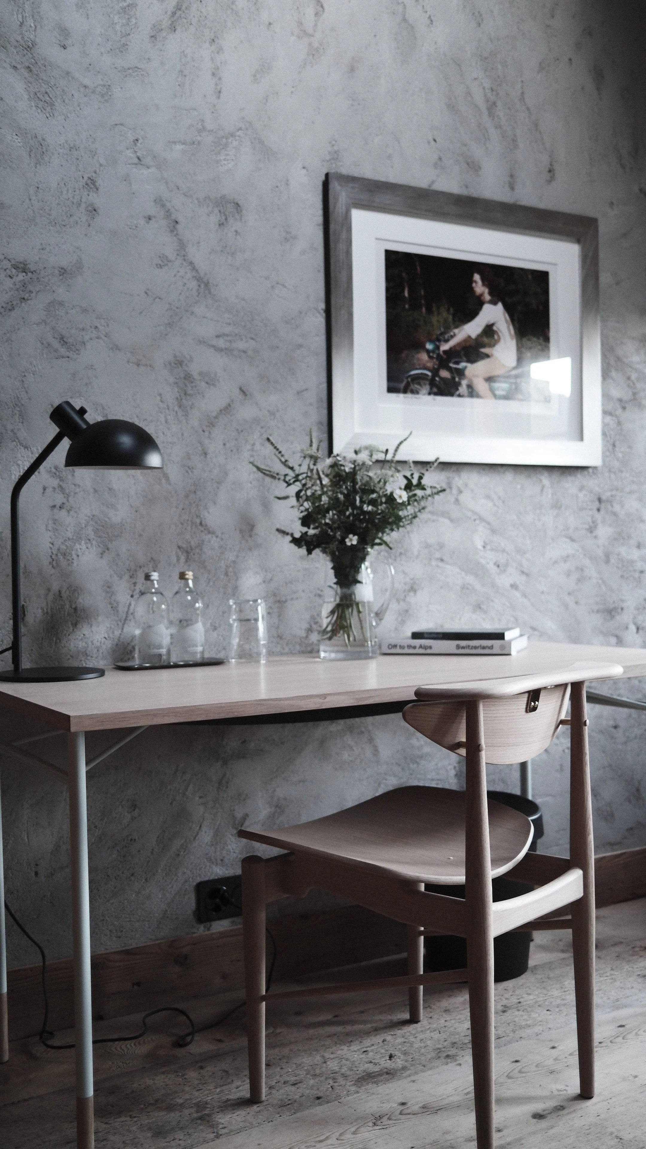 A modern home office corner with a wooden desk, a wooden chair, a black desk lamp, and decorative items including a vase of flowers, bottled water, a glass, and books on a textured gray wall background.