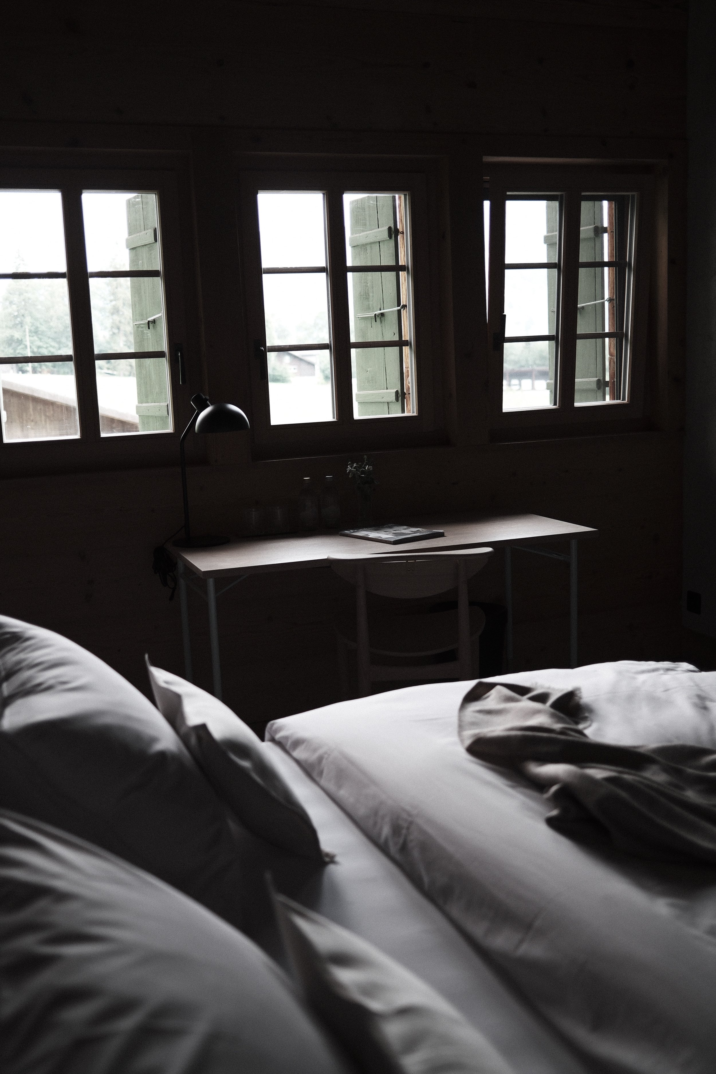 A bedroom with a row of three windows with open green shutters, a desk with a black desk lamp, and a bed with white sheets in the foreground.