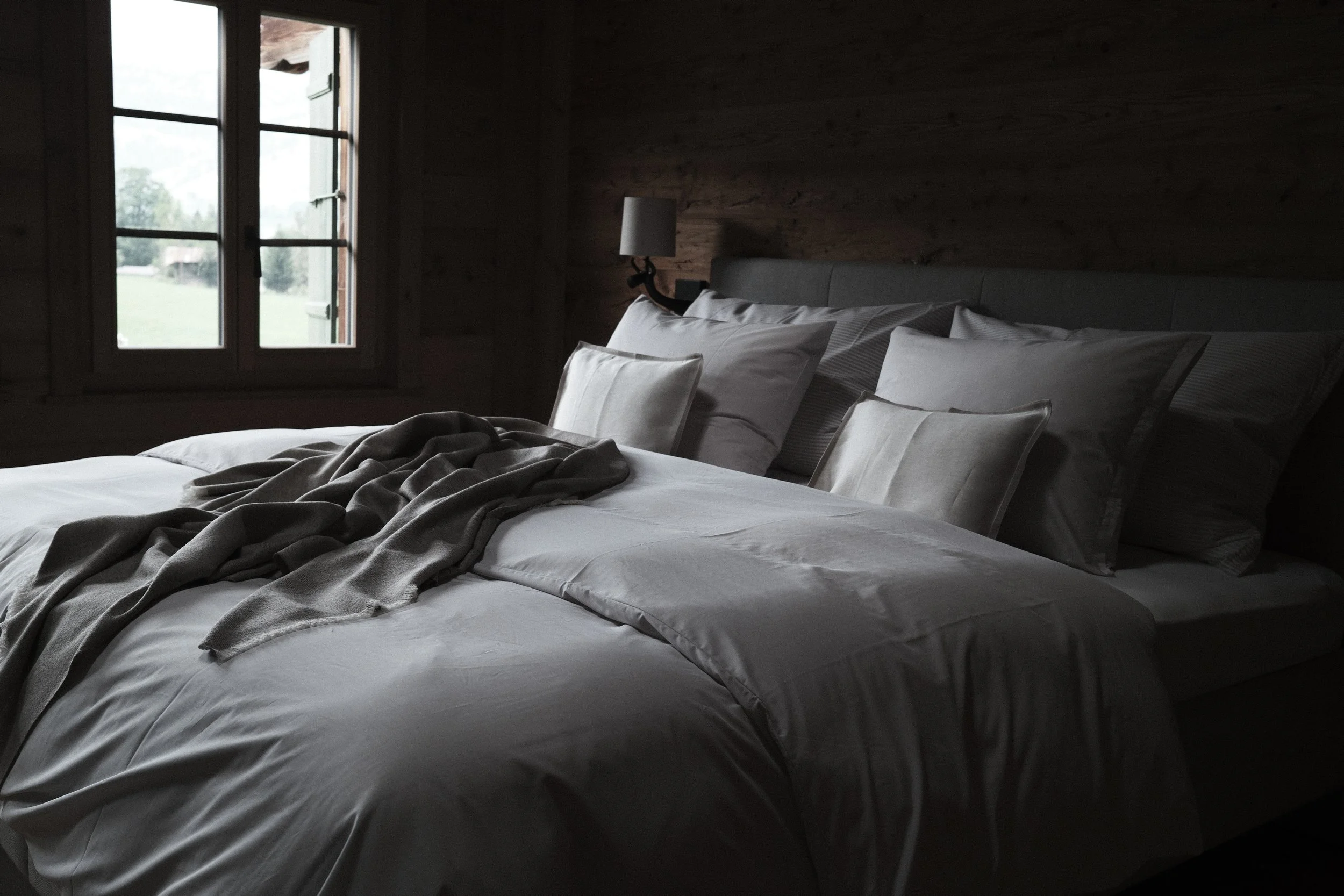 A bedroom with a bed covered in white sheets and multiple pillows, a blanket draped at the foot of the bed, a window showing an outdoor view of trees and grass, and a wooden wall with a small wall-mounted lamp beside the bed.