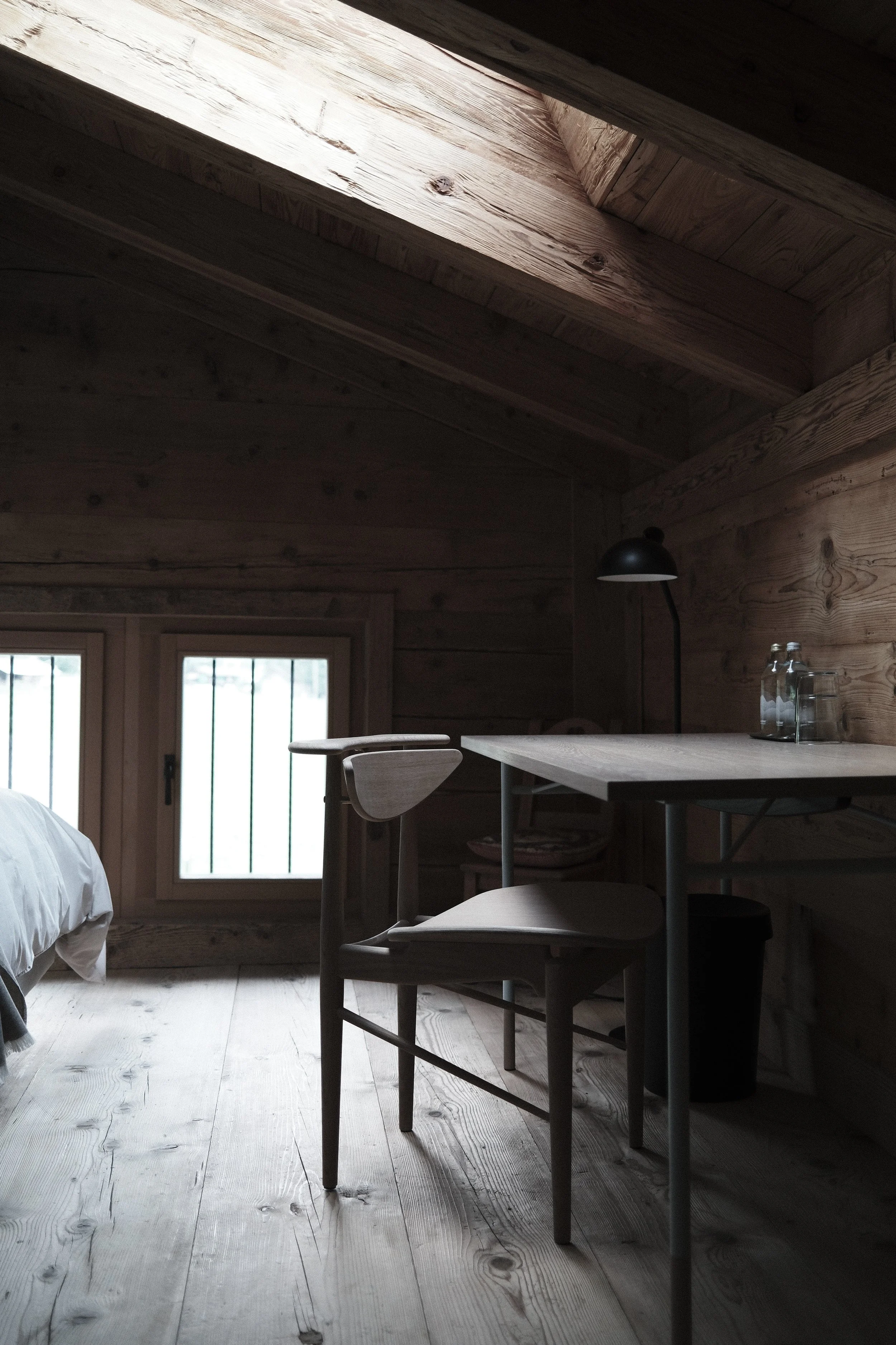 Interior of a cozy wooden cabin with a sloped ceiling and a large window, featuring a dining table, chairs, a black wall-mounted lamp, and a bed in the corner.