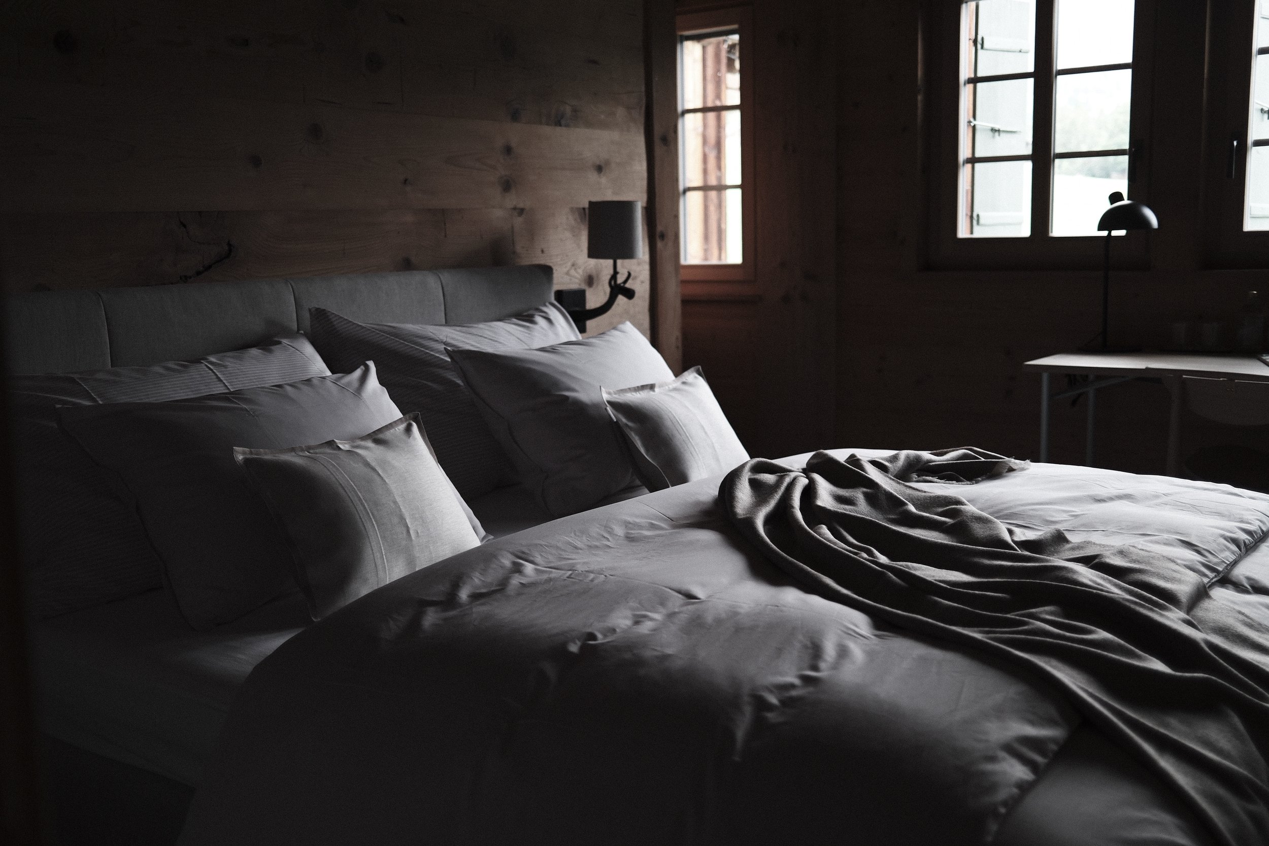 Dimly lit bedroom with a bed covered in white sheets and pillows, a blanket draped over the bed, wooden wall paneling, a nightstand with a black lamp, and two windows with open shutters allowing natural light.