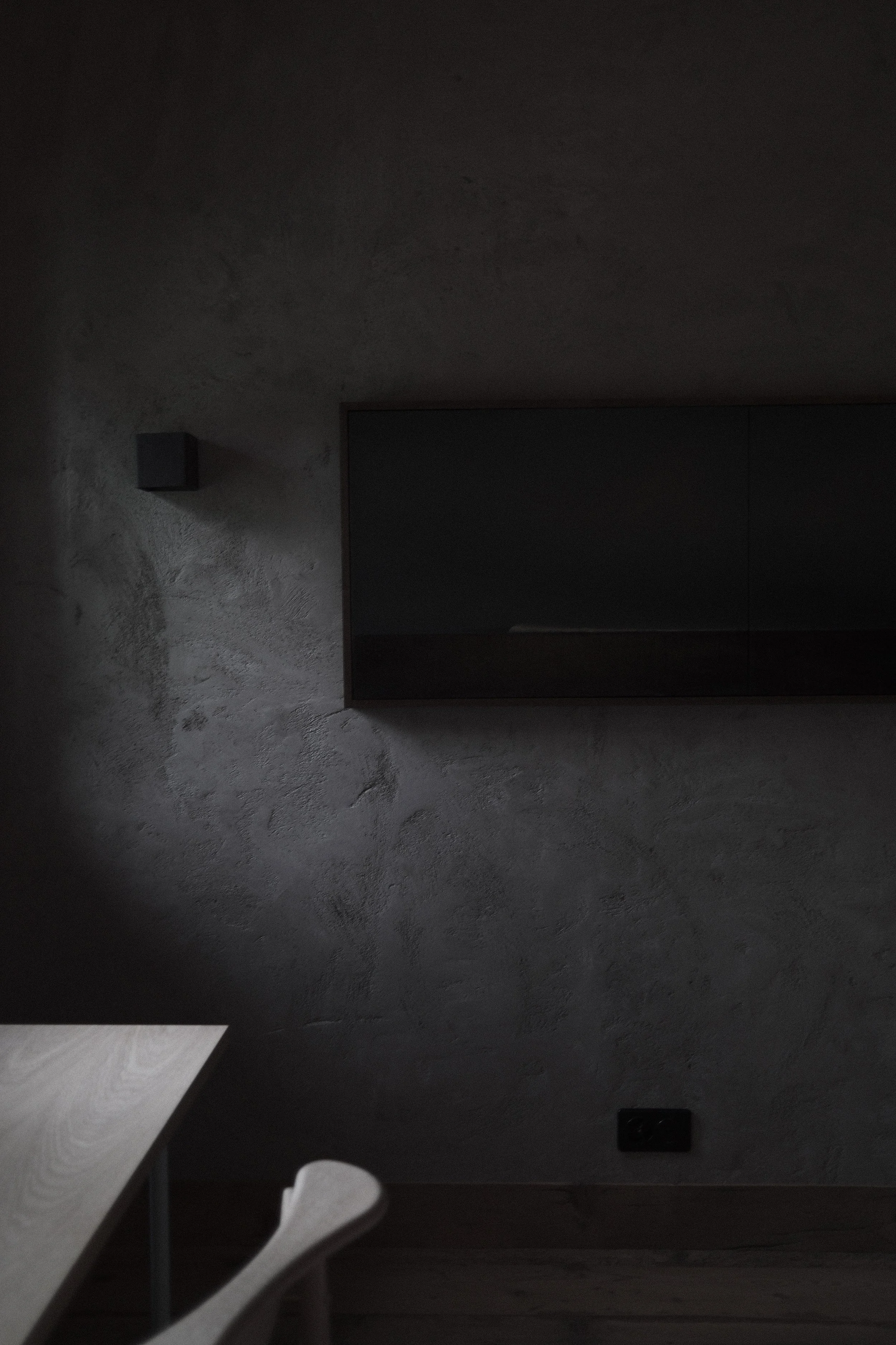 A dark, textured wall with a small black box on it. A mirror or glass surface is partially visible, reflecting some light. Part of a white object, possibly a chair or furniture, is visible in the lower left corner.