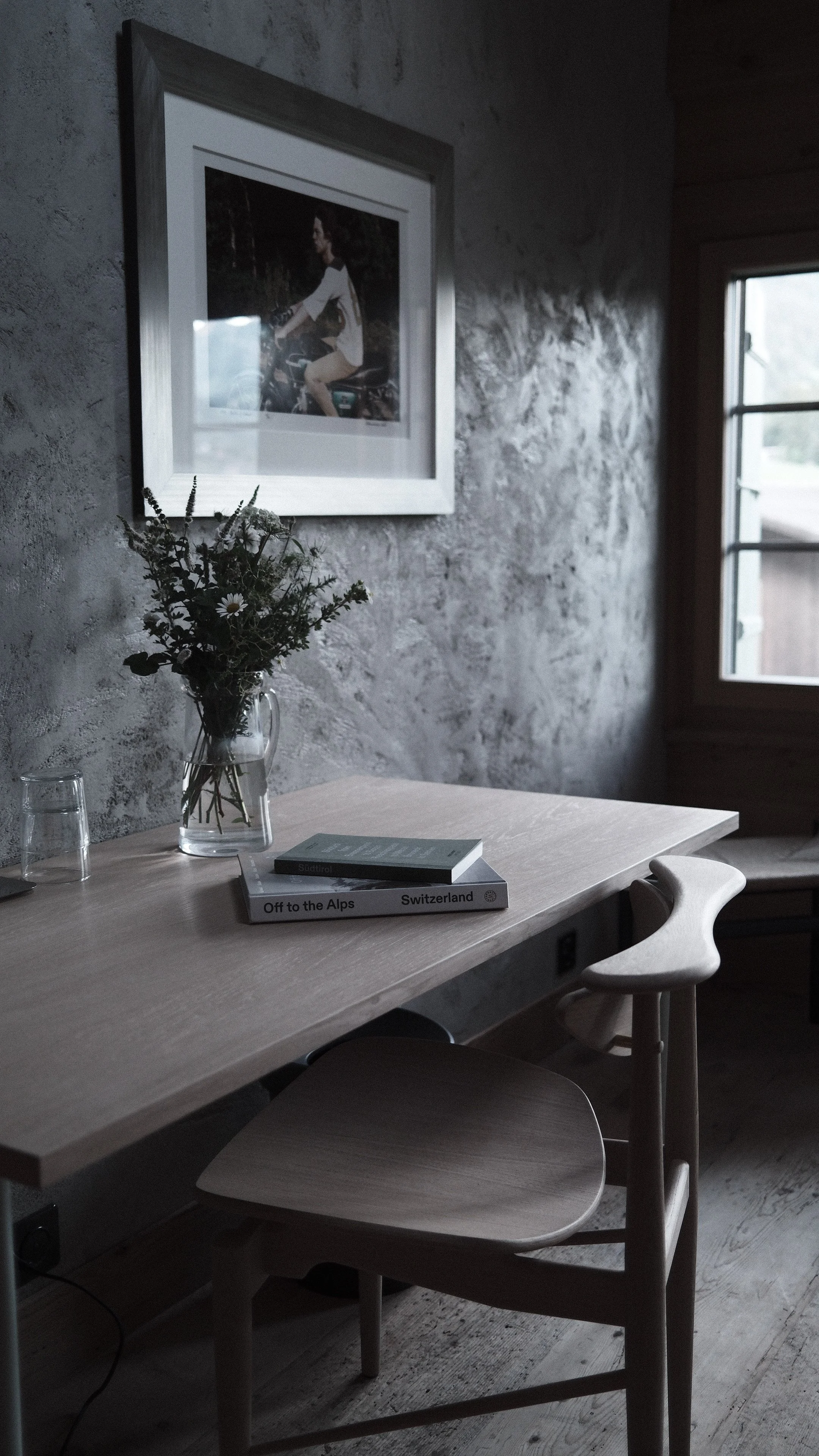 A minimalist wooden desk with a glass vase of wildflowers, two stacked books titled 'Off to the Alps' and 'Switzerland', and a glass of water. Behind the desk is a white chair and a textured concrete wall with a framed photograph of a woman riding a motorcycle. A window with wooden trim is on the right side, allowing natural light into the room.