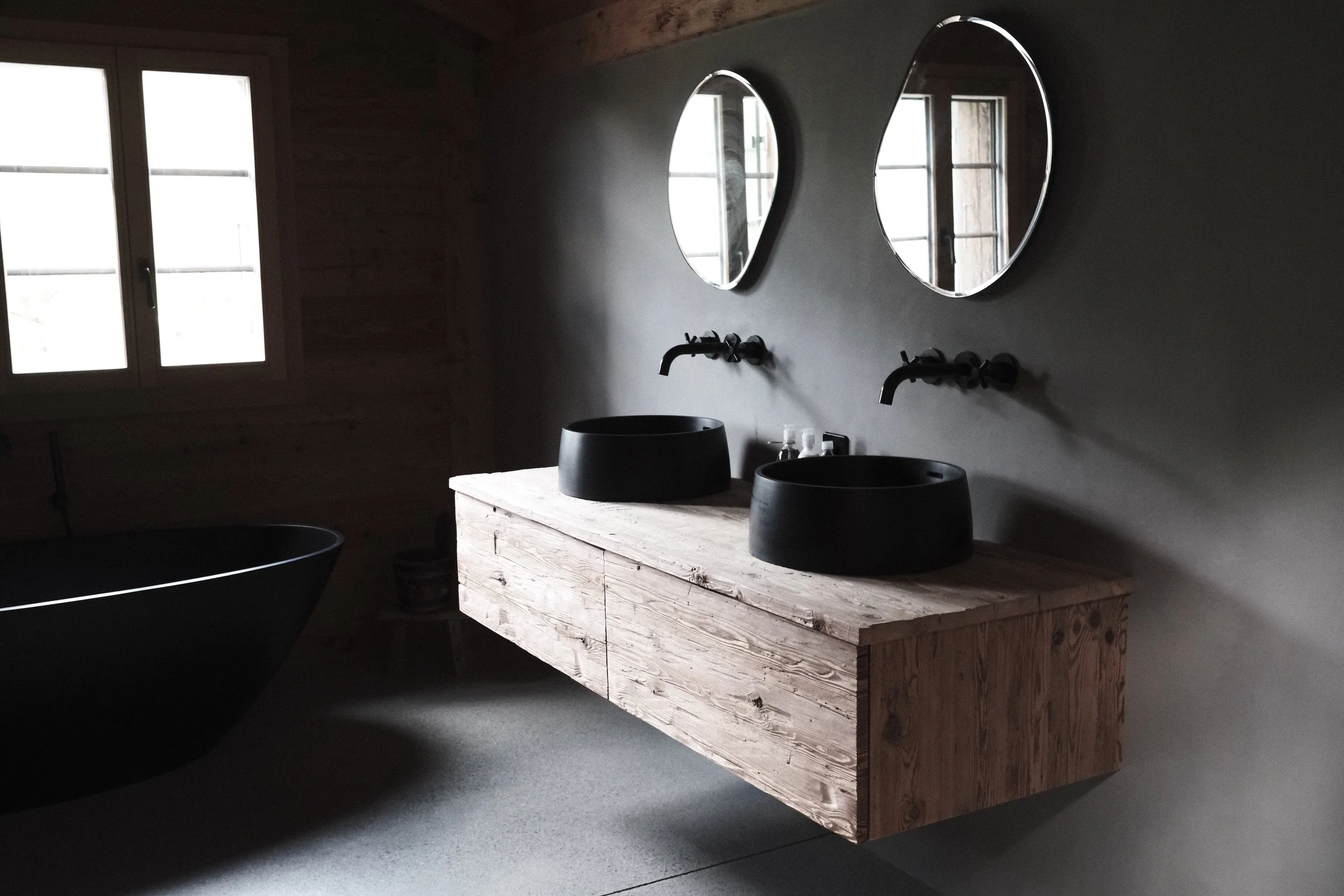 A rustic bathroom vanity made of unfinished wood with two black vessel sinks and wall-mounted black faucets, two round mirrors above, and a window to the left letting in light.