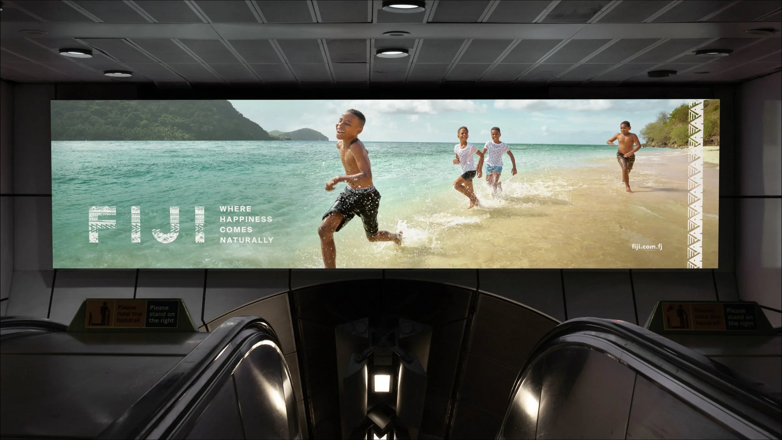 Billboard advertisement showing children playing in the ocean on a beach, with the slogan 'Where happiness comes naturally' and a website link 'fiji.com.fj'.
