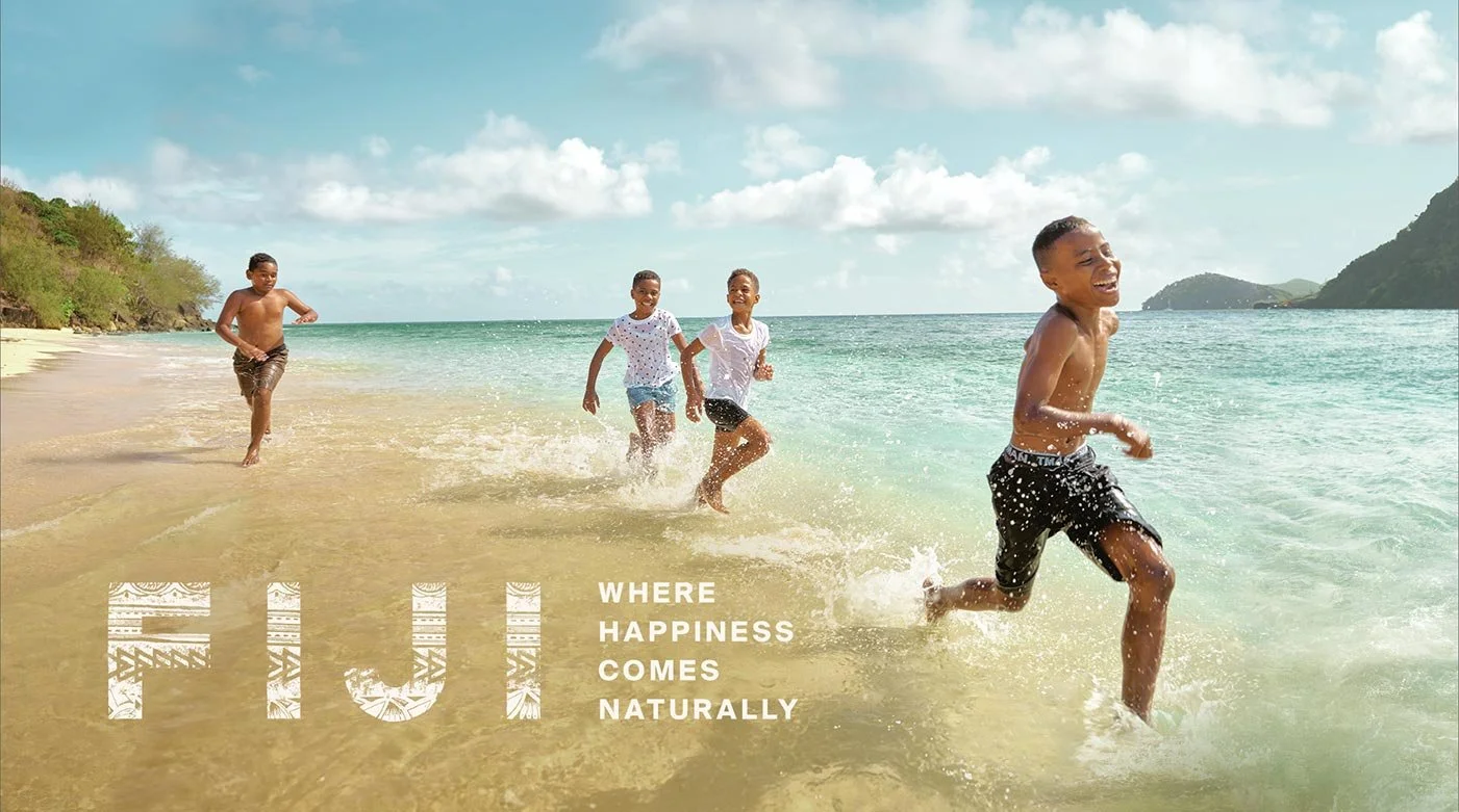 Four children playing and running in the shallow water at the beach, smiling and enjoying sunny weather with a scenic background of hills, sand, and clouds. Overlaid text reads 'WHERE HAPPINESS COMES NATURALLY'.