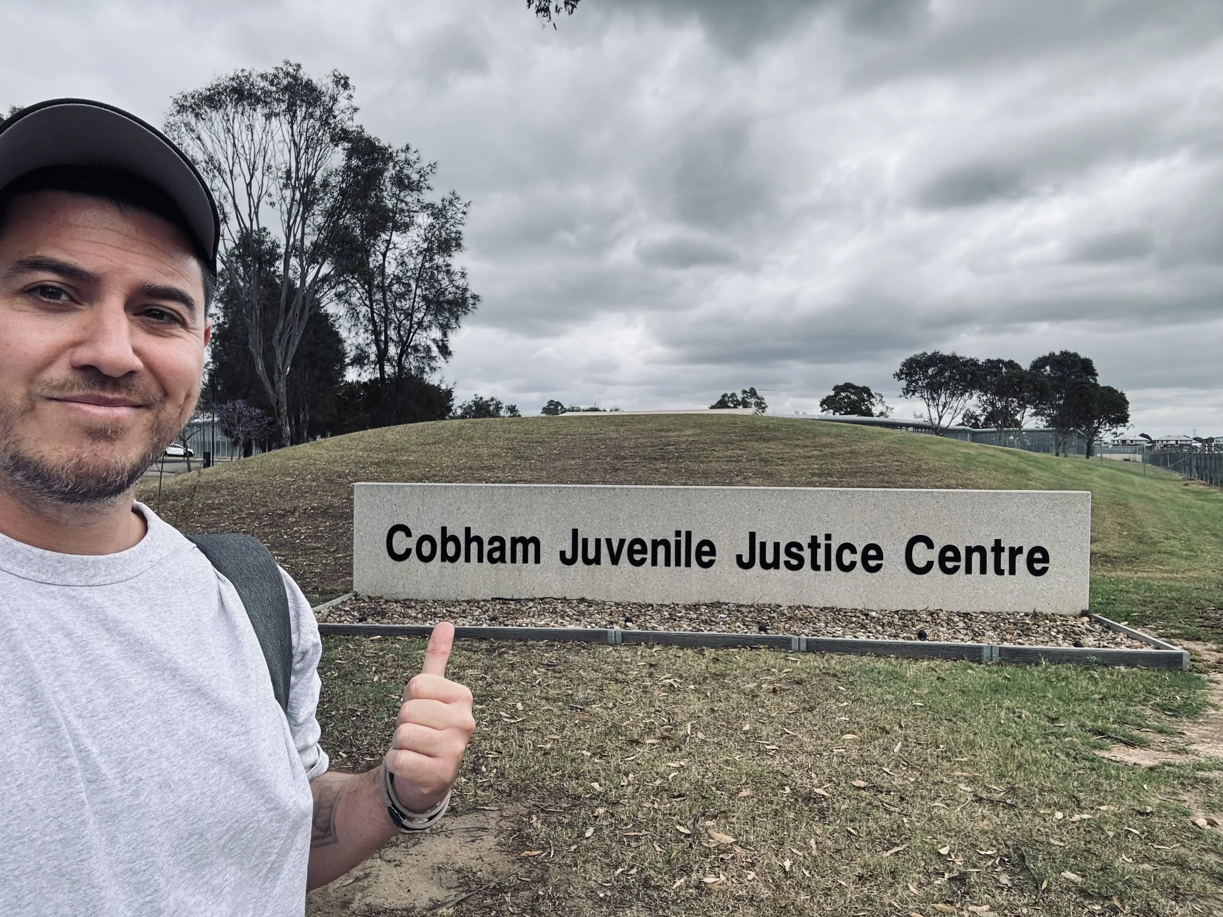 A man with a gray t-shirt and gray cap standing outdoors, pointing at a sign that says 'Cobham Juvenile Justice Centre'. The background features trees, cloudy sky, and a small grassy hill.